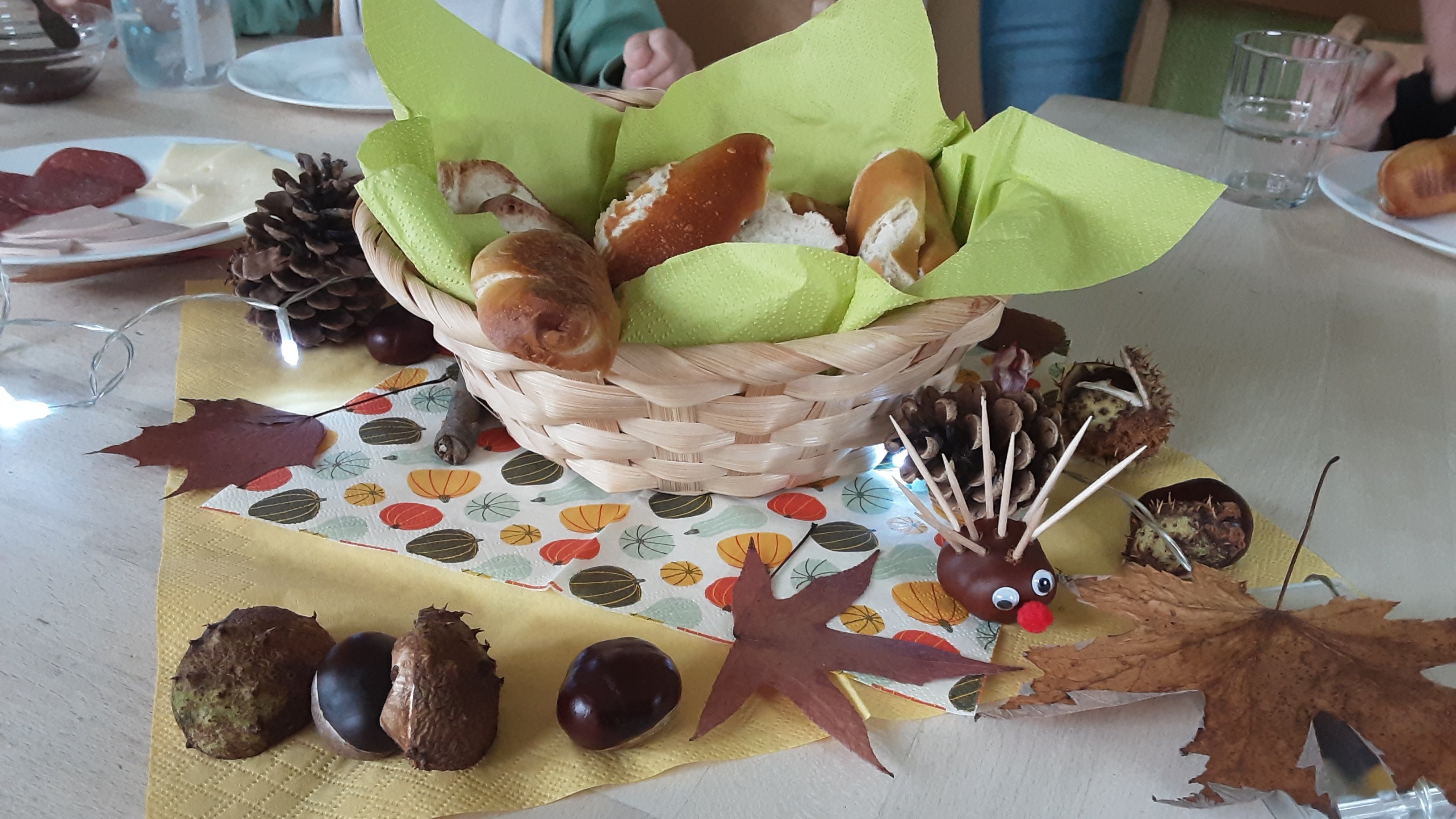 Auf dem Foto ist eine herbstlich dekorierte Tischszene zu sehen, wahrscheinlich im Rahmen eines Herbstfests oder Frühstücks in einer Kita/Einrichtung. Hier die Details: Tischdekoration: Im Zentrum steht ein kleiner Brotkorb aus hellem Flechtwerk, gefüllt mit verschiedenen Brötchen (u.a. Laugengebäck). Der Korb ist mit einer hellgrünen Serviette ausgelegt. Rund um den Korb und auf einer gelb-gemusterten Tischdecke/Serviette liegen verschiedene Herbstmaterialien: Kastanien (glänzende, braune sowie noch in ihrer stacheligen Schale) ? Tannenzapfen ? Trockene Laubblätter (braun und rotbraun) ? Ein kleiner Kastanienigel mit Wackelaugen und roter Nase ist ebenfalls zu sehen. ? Eine Lichterkette (mit kleinen LEDs) ist dezent in die Dekoration eingearbeitet. ✨ Geschirr/Essen im Hintergrund: Auf einem Teller links liegen Aufschnitt und Käse (Wurst und Käsescheiben). ?? Man sieht weiße Teller, Gläser und Besteck, was auf eine Mahlzeit hindeutet. Personen (angedeutet): Im Hintergrund sind verschwommen die Hände und die Kleidung von mindestens zwei Personen zu erkennen, die am Tisch sitzen. Insgesamt zeigt das Bild eine gemütliche, herbstliche Mahlzeit mit viel Liebe zum Detail in der Dekoration.