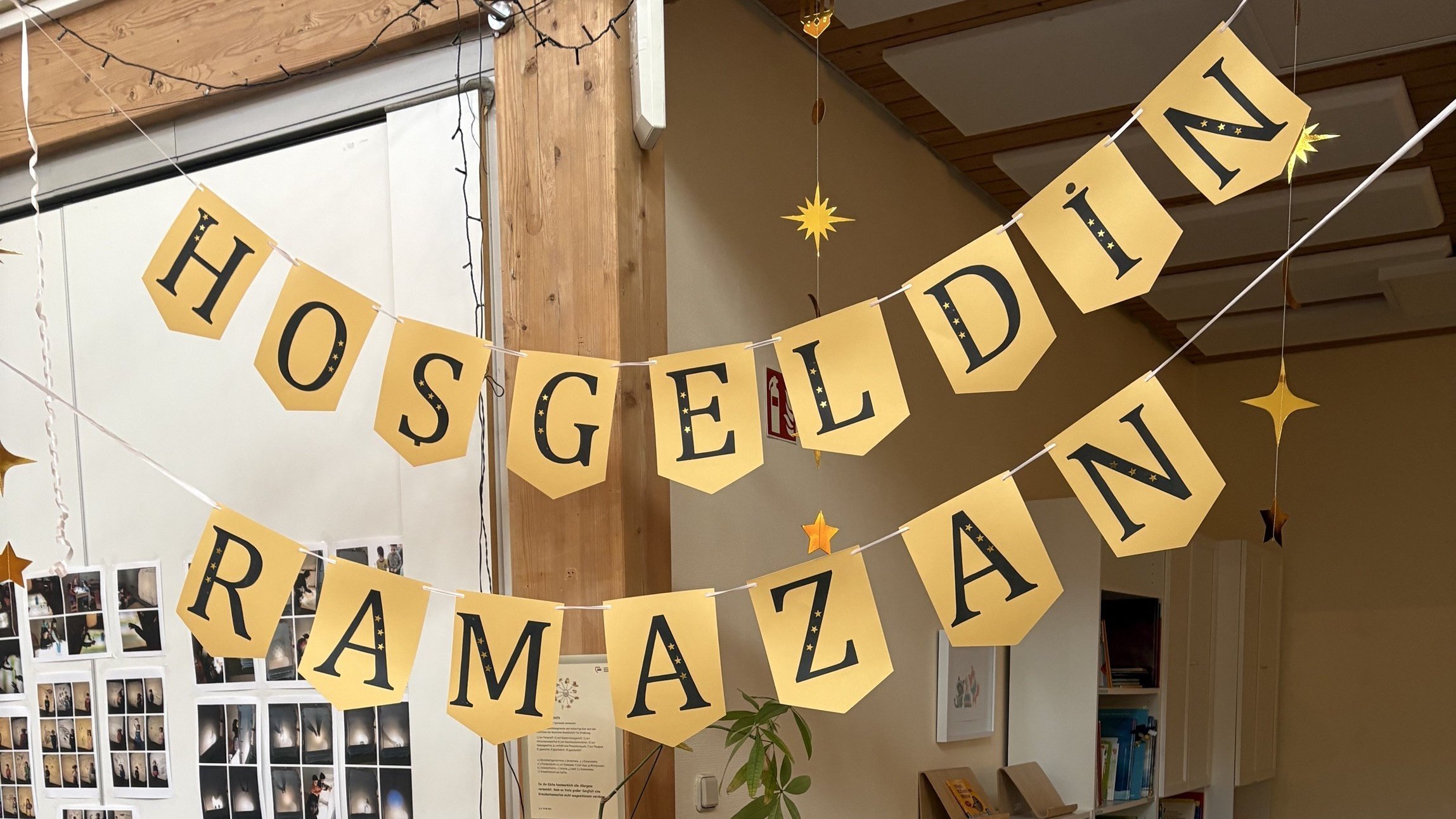 Auf dem Foto sieht man eine liebevoll dekorierte Ecke in einer Kita oder ähnlichen Einrichtung zum Beginn des Ramadan. Oben hängen zwei gelbe Girlanden mit der Aufschrift „HOSGELDIN RAMAZAN“ (Willkommen Ramadan). Auf der Theke stehen in einer Reihe bunte Tütchen mit Zahlen – ähnlich wie ein Adventskalender, nur für den Ramadan. An der Vorderseite des Tresens ist ein gelbes Schild mit Lichterkette befestigt, auf dem mehrsprachig „Willkommen Ramadan / Marhaban ya Ramadan / Welcome Ramadan“ steht, umgeben von Monden, Sternen und Laternen. Links sind außerdem Bildleisten mit Symbolkarten für den Wochenplan (Mi, Do, Fr) zu sehen.