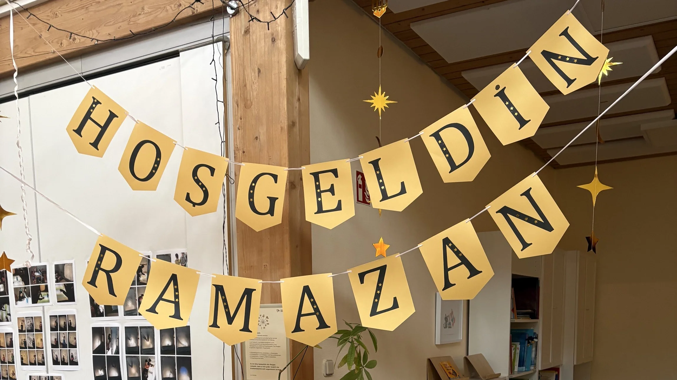 Auf dem Foto sieht man eine liebevoll dekorierte Ecke in einer Kita oder ähnlichen Einrichtung zum Beginn des Ramadan. Oben hängen zwei gelbe Girlanden mit der Aufschrift „HOSGELDIN RAMAZAN“ (Willkommen Ramadan). Auf der Theke stehen in einer Reihe bunte Tütchen mit Zahlen – ähnlich wie ein Adventskalender, nur für den Ramadan. An der Vorderseite des Tresens ist ein gelbes Schild mit Lichterkette befestigt, auf dem mehrsprachig „Willkommen Ramadan / Marhaban ya Ramadan / Welcome Ramadan“ steht, umgeben von Monden, Sternen und Laternen. Links sind außerdem Bildleisten mit Symbolkarten für den Wochenplan (Mi, Do, Fr) zu sehen.