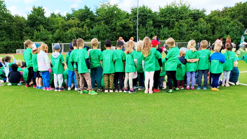 Auf dem Foto ist eine große Gruppe von Kindern zu sehen, die auf einem Sportplatz oder Fußballfeld stehen. Die Kinder tragen grüne T-Shirts mit weißem Aufdruck auf dem Rücken – vermutlich einheitliche Shirts für eine Veranstaltung. Die Kinder stehen mit dem Rücken zur Kamera, viele in einer Reihe, einige sitzen oder knien am Boden. Es sieht nach einem Gruppenereignis oder einer sportlichen Aktivität aus, möglicherweise einem Spendenlauf oder Sporttag für Kinder. Im Hintergrund sind Erwachsene und Spielgeräte oder eine Hüpfburg zu sehen, was auf eine organisierte Veranstaltung mit mehreren Aktivitäten hinweist.