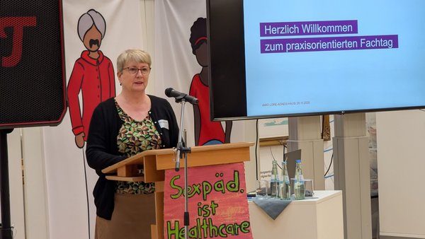 Auf dem Bild ist eine Veranstaltung oder Konferenz zu sehen. Hier sind die Details: Vordergrund: Eine Frau steht an einem Holzpult und spricht in ein Mikrofon. Sie trägt eine Brille, ein gemustertes Oberteil und eine dunkle Strickjacke. Pult-Dekoration: Am Pult hängt ein Schild, auf dem in bunter Schrift steht: "Sexpäd. ist Healthcare" (was wahrscheinlich "Sexualpädagogik ist Gesundheitsfürsorge" bedeutet). Hintergrund (Leinwand/Bildschirm): Ein großer Bildschirm zeigt eine Präsentation mit dem Text: "Herzlich Willkommen zum praxisorientierten Fachtag" Darunter steht das Logo der AWO (Arbeiterwohlfahrt) und der Text "Beratungszentrum Lippe/OWL" sowie das Datum "AWO JULE AUBI | 26.11.2025". Weitere Hintergründelemente: Links hinter der Rednerin stehen Banner mit stilisierten Illustrationen von Menschen. Ganz links ist ein Lautsprecher zu erkennen. Rechts neben dem Pult steht ein kleiner Tisch mit Wasserflaschen und Gläsern.