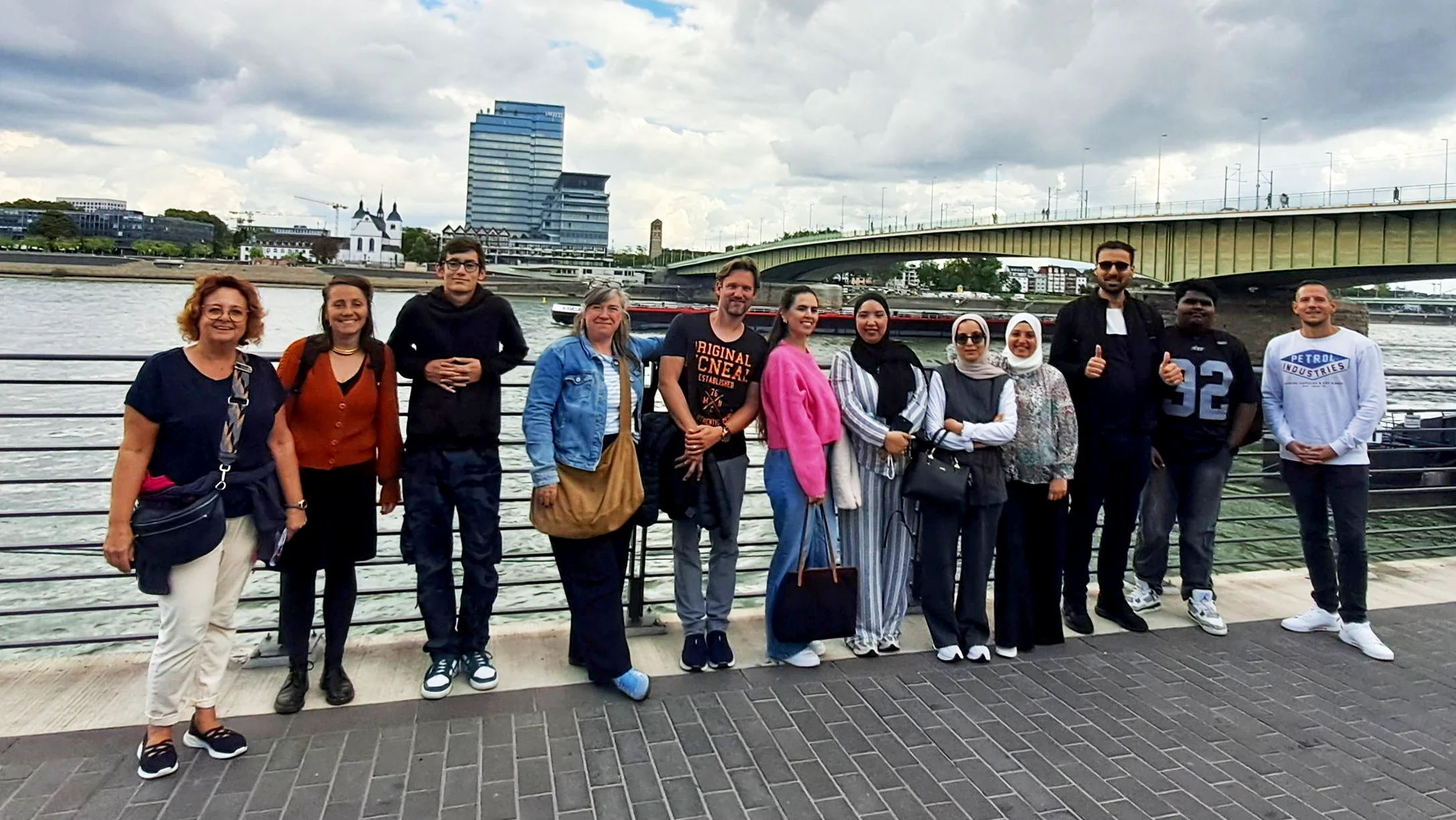Auf dem Foto ist eine Gruppe von 13 Erwachsenen zu sehen, die auf einem gepflasterten Uferweg am Wasser stehen. Im Hintergrund sind ein Fluss, eine Brücke und moderne Gebäude zu erkennen. Der Himmel ist bewölkt.