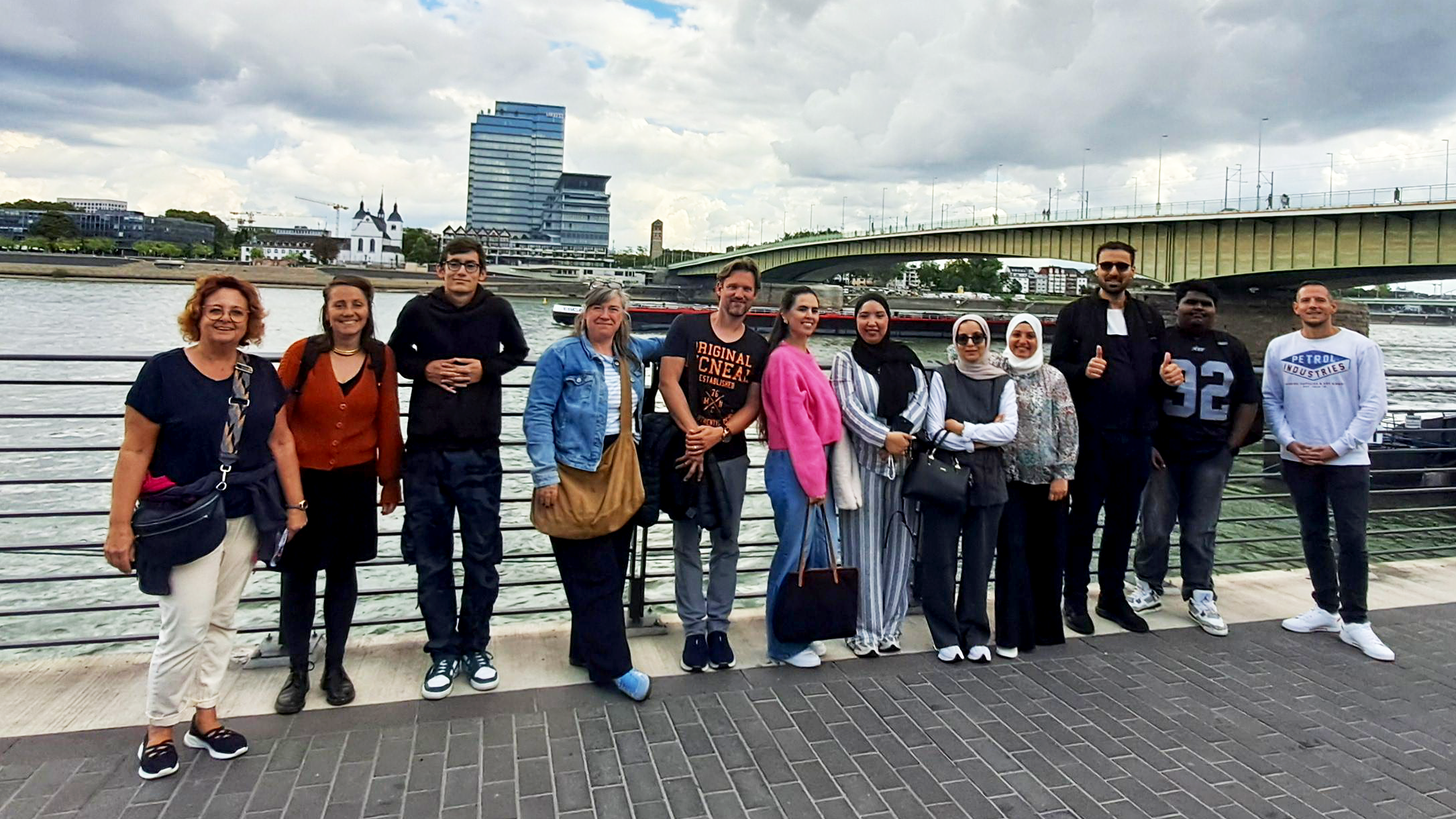 Auf dem Foto ist eine Gruppe von 13 Erwachsenen zu sehen, die auf einem gepflasterten Uferweg am Wasser stehen. Im Hintergrund sind ein Fluss, eine Brücke und moderne Gebäude zu erkennen. Der Himmel ist bewölkt.