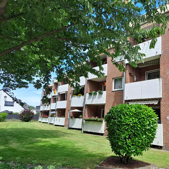 Auf dem Foto ist ebenfalls ein mehrstöckiges Wohngebäude zu sehen, vermutlich ein Mehrfamilienhaus. Das Gebäude besteht aus rotem Backstein und hat mehrere weiße Balkone, die zum Teil mit Blumen und Pflanzen geschmückt sind. Im Vordergrund sieht man eine gepflegte Grünfläche mit einem kleinen, kugelrund geschnittenen Strauch und einem gepflasterten Gehweg. Links im Bild steht ein großer Laubbaum, dessen Blätter teilweise den Blick auf das Gebäude überdecken und Schatten spenden. Im Hintergrund links sind weitere Wohnhäuser zu erkennen. Auch dieses Bild wurde bei sonnigem Wetter mit blauem Himmel aufgenommen. Die Szene wirkt ruhig, ordentlich und gepflegt.