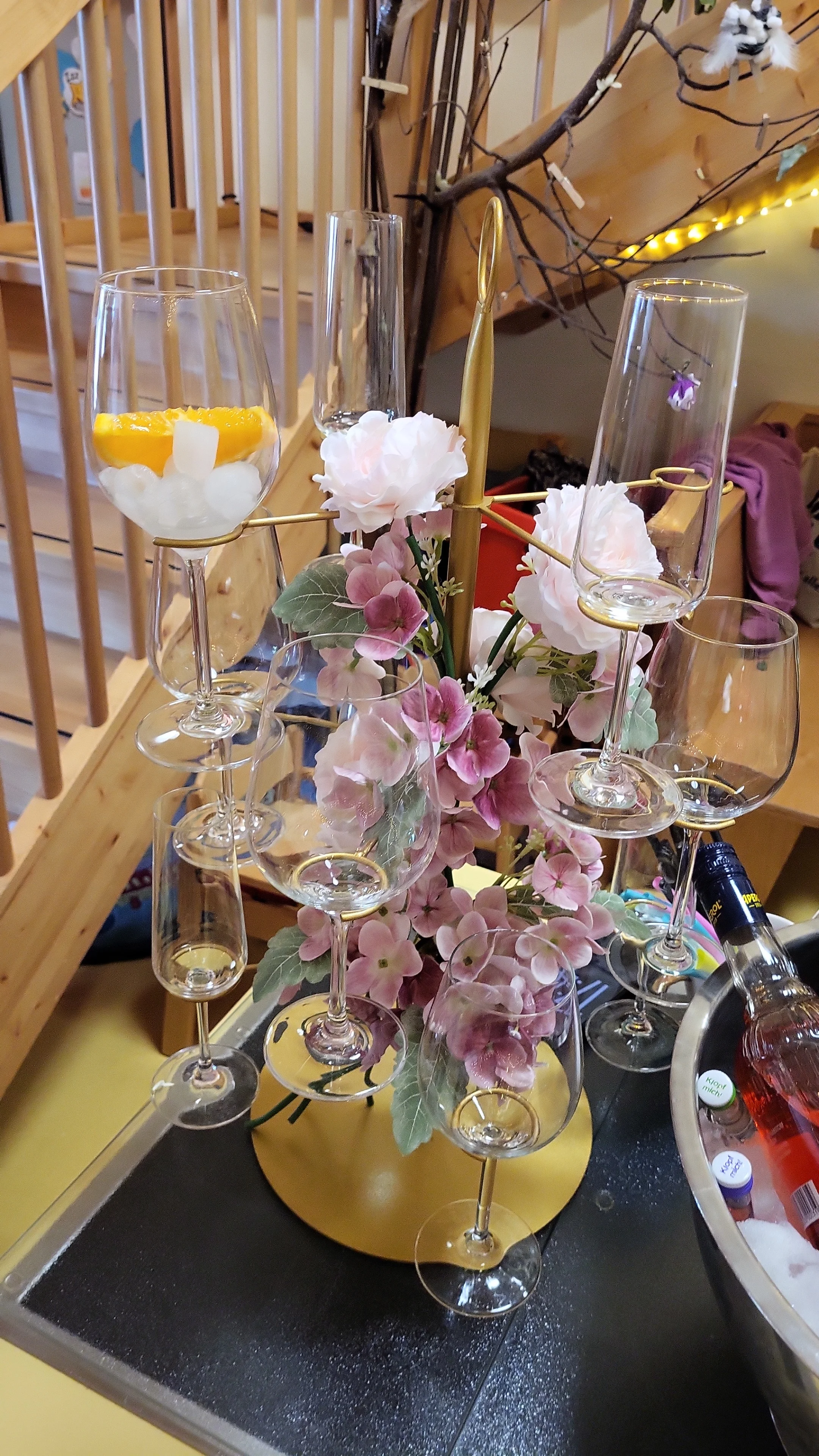 Auf dem Bild ist ein dekorativ arrangierter Glasständer zu sehen, auf dem mehrere leere Wein- und Sektgläser hängen bzw. stehen. In der Mitte sind künstliche rosa und weiße Blumen angebracht, daneben erkennt man einen Sekt-/Getränkekühler mit Flaschen, im Hintergrund ein Holztreppengeländer und etwas Deko (Zweige, Lichterkette).
