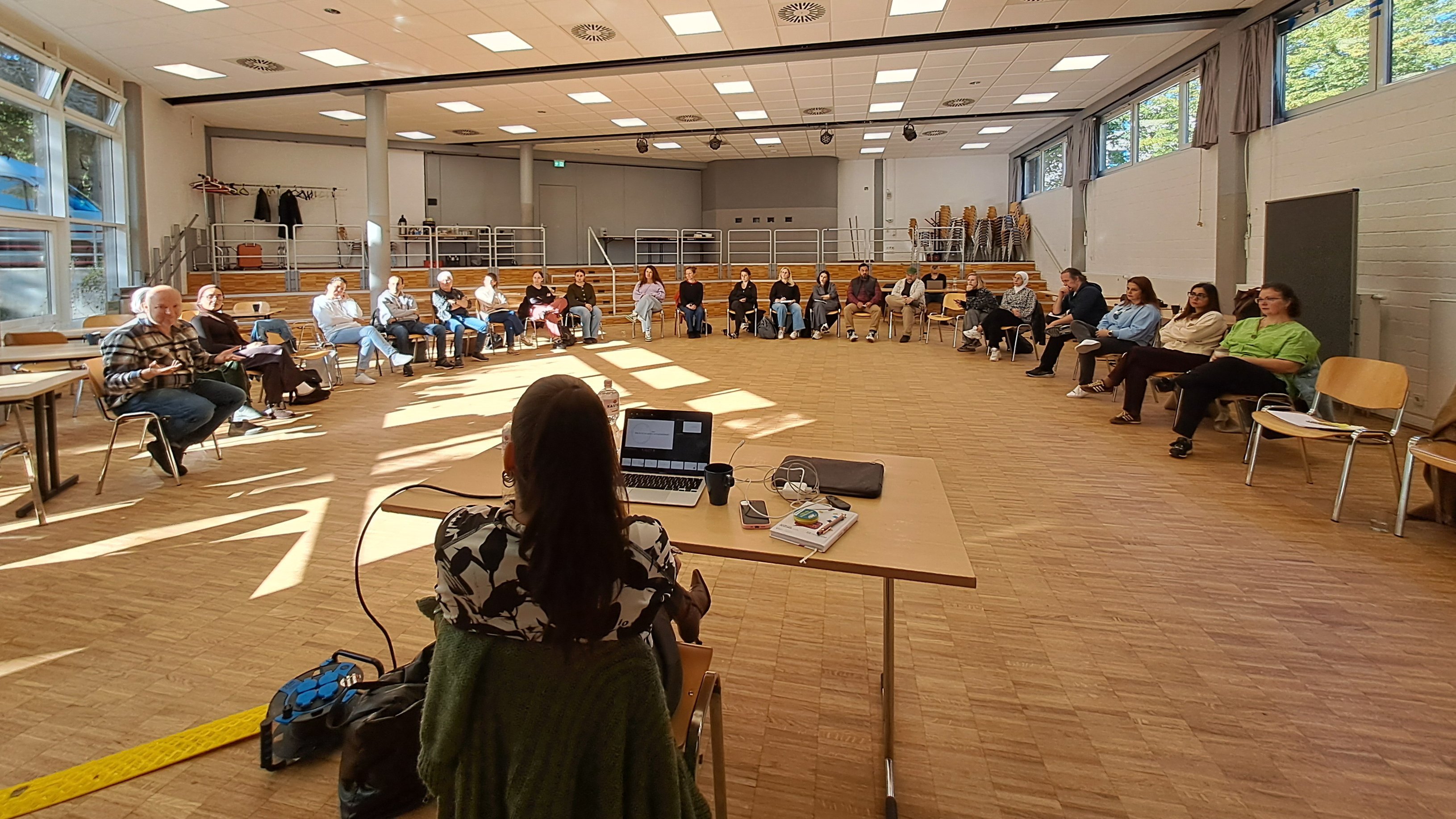  Auf dem Foto ist eine Gruppe von Erwachsenen zu sehen, die an einer Versammlung, Schulung oder einem Workshop in einem großen Raum teilnehmen. Hier sind die Details: Personen und Anordnung: Etwa 20 bis 25 Personen sitzen in einem großen, offenen Raum. Sie sind in einem Halbkreis auf Stühlen angeordnet und schauen in Richtung des vorderen Bereichs. Eine weibliche Person sitzt vorne an einem Tisch mit einem Laptop und ist dem Publikum zugewandt (ihr Rücken ist zur Kamera). Sie scheint die Moderatorin oder Vortragende zu sein. Raum: Es handelt sich um einen großen Mehrzweckraum oder eine Aula mit hellem Holzfußboden. Der Raum hat große Fenster an den Seiten, durch die viel natürliches Licht einfällt. Man sieht Bäume und blauen Himmel draußen. ☀️ Die Decke besteht aus vielen quadratischen Lichtelementen (Rasterdecke). Es gibt eine Bühnentechnik mit Strahlern und einem Beamer/Projektor (der auf eine nicht sichtbare Leinwand projiziert). Im Hintergrund gibt es eine erhöhte Tribüne oder Sitzreihe und gestapelte Stühle, was auf die Nutzung als Versammlungsraum hindeutet. Zusammenfassend zeigt das Bild eine organisierte Veranstaltung in einem großen, hellen Raum.