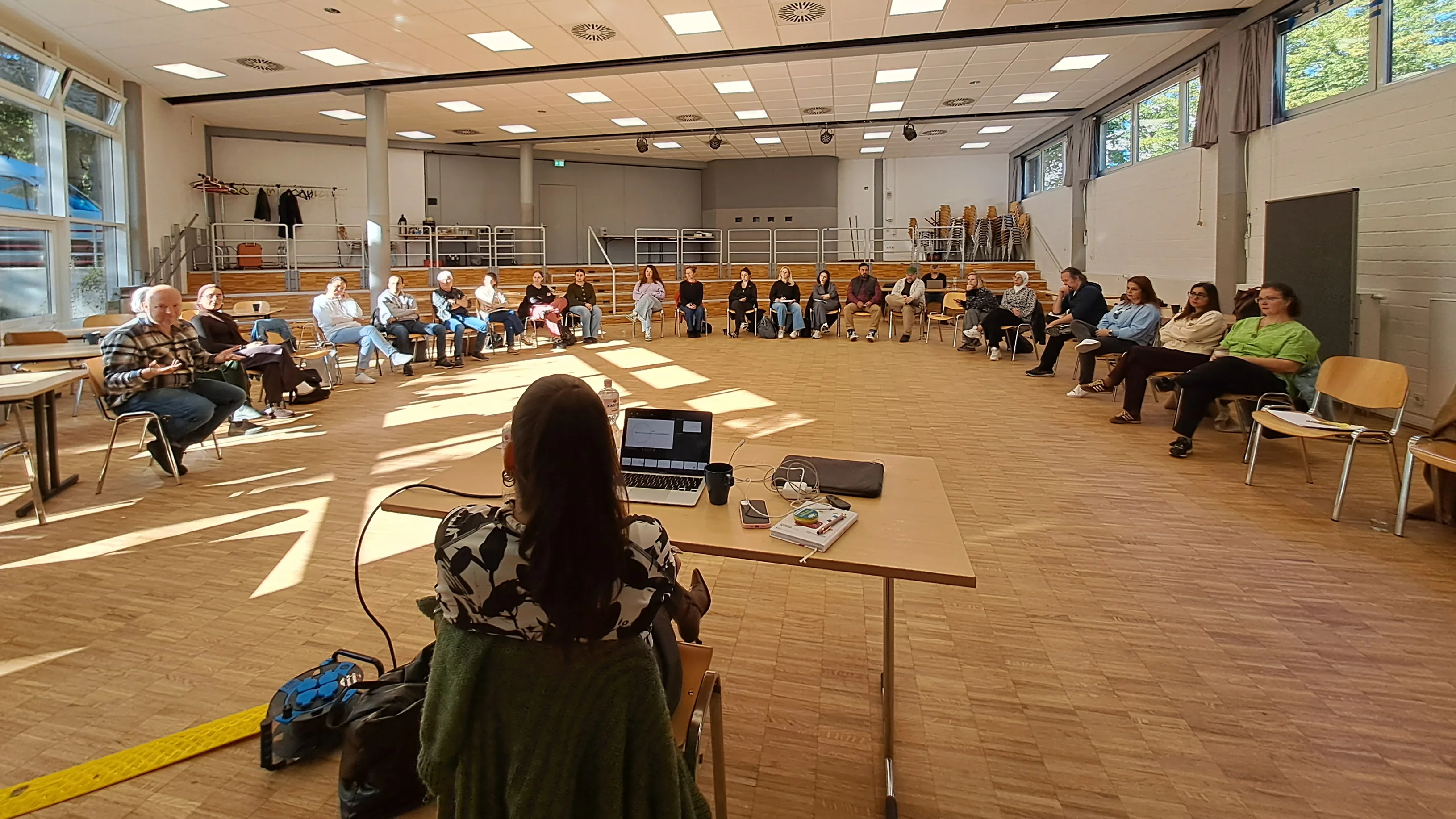  Auf dem Foto ist eine Gruppe von Erwachsenen zu sehen, die an einer Versammlung, Schulung oder einem Workshop in einem großen Raum teilnehmen. Hier sind die Details: Personen und Anordnung: Etwa 20 bis 25 Personen sitzen in einem großen, offenen Raum. Sie sind in einem Halbkreis auf Stühlen angeordnet und schauen in Richtung des vorderen Bereichs. Eine weibliche Person sitzt vorne an einem Tisch mit einem Laptop und ist dem Publikum zugewandt (ihr Rücken ist zur Kamera). Sie scheint die Moderatorin oder Vortragende zu sein. Raum: Es handelt sich um einen großen Mehrzweckraum oder eine Aula mit hellem Holzfußboden. Der Raum hat große Fenster an den Seiten, durch die viel natürliches Licht einfällt. Man sieht Bäume und blauen Himmel draußen. ☀️ Die Decke besteht aus vielen quadratischen Lichtelementen (Rasterdecke). Es gibt eine Bühnentechnik mit Strahlern und einem Beamer/Projektor (der auf eine nicht sichtbare Leinwand projiziert). Im Hintergrund gibt es eine erhöhte Tribüne oder Sitzreihe und gestapelte Stühle, was auf die Nutzung als Versammlungsraum hindeutet. Zusammenfassend zeigt das Bild eine organisierte Veranstaltung in einem großen, hellen Raum.