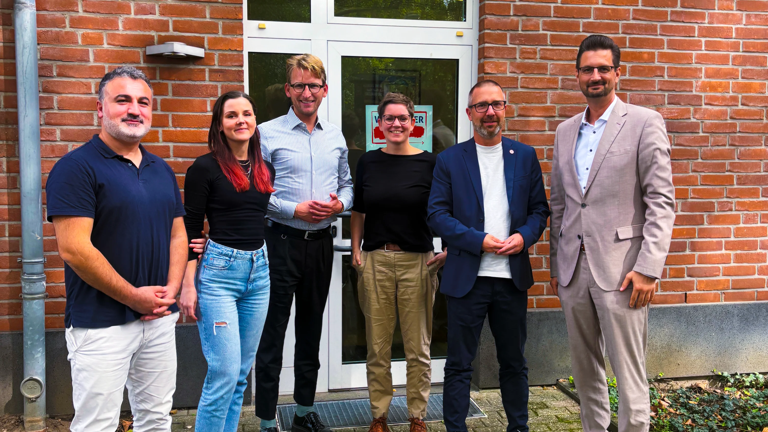 Auf dem Foto stehen sechs Personen vor dem Eingang eines Backsteingebäudes. Sie sind offenbar für ein gemeinsames Gruppen- bzw. Pressfoto zusammengekommen. Im Hintergrund sieht man eine Glastür, einen gelb verputzten Fassadenabschnitt, ein Fallrohr und einen gepflasterten Gehweg. Die Gruppe ist unterschiedlich formell gekleidet – von Polohemd und Jeans bis zu Sakko und hellem Anzug. Auf dem Schild in der Tür ist teilweise „…CENTER“ zu erkennen; der restliche Text ist nicht eindeutig lesbar.