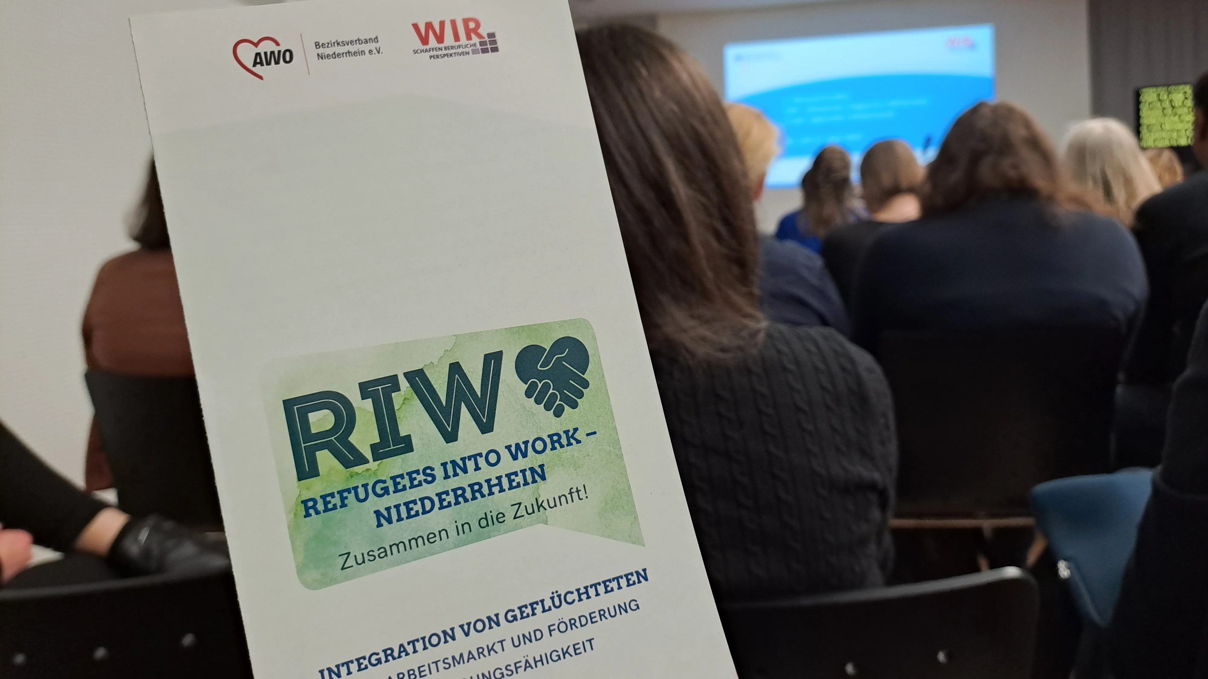 Auf dem Foto ist eine Veranstaltung oder ein Vortrag zu sehen, der sich mit dem Thema Integration von Geflüchteten in den Arbeitsmarkt beschäftigt. Im Vordergrund hält eine Person einen Flyer oder ein Faltblatt in der Hand. Darauf steht: „RIW – Refugees into Work – Niederrhein. Zusammen in die Zukunft!“ „Integration von Geflüchteten in den Arbeitsmarkt und Förderung der Beschäftigungsfähigkeit“ Oben auf dem Flyer sind die Logos von zwei Organisationen zu sehen: AWO Bezirksverband Niederrhein e.V. WIR – Schaffen berufliche Perspektiven Im Hintergrund erkennt man ein Publikum, das auf eine Präsentation blickt, die auf einer Leinwand vorne im Raum gezeigt wird.
