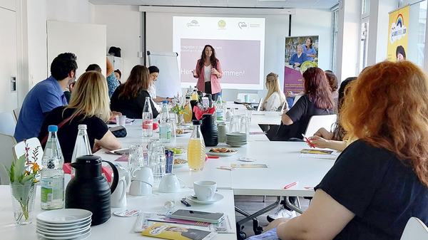 Auf dem Foto ist eine Veranstaltung oder ein Workshop in einem Konferenzraum zu sehen. Die Atmosphäre wirkt professionell und einladend. Mehrere Menschen sitzen an einem langen Tisch und hören aufmerksam einer Frau zu, die vorne steht und eine Präsentation hält. Details: • Auf dem Bildschirm steht: „Herzlich willkommen!“, sowie „Integration in den Arbeitsmarkt – Komm an in der Pflege“. Dies deutet auf eine Veranstaltung zur Arbeitsmarktintegration hin. • Es sind Logos der AWO und von INAR zu sehen. • Auf dem Tisch stehen Wasserflaschen, Kaffee, Saft, Snacks und Informationsmaterialien. • Im Hintergrund hängen Roll-Ups von der AWO.
