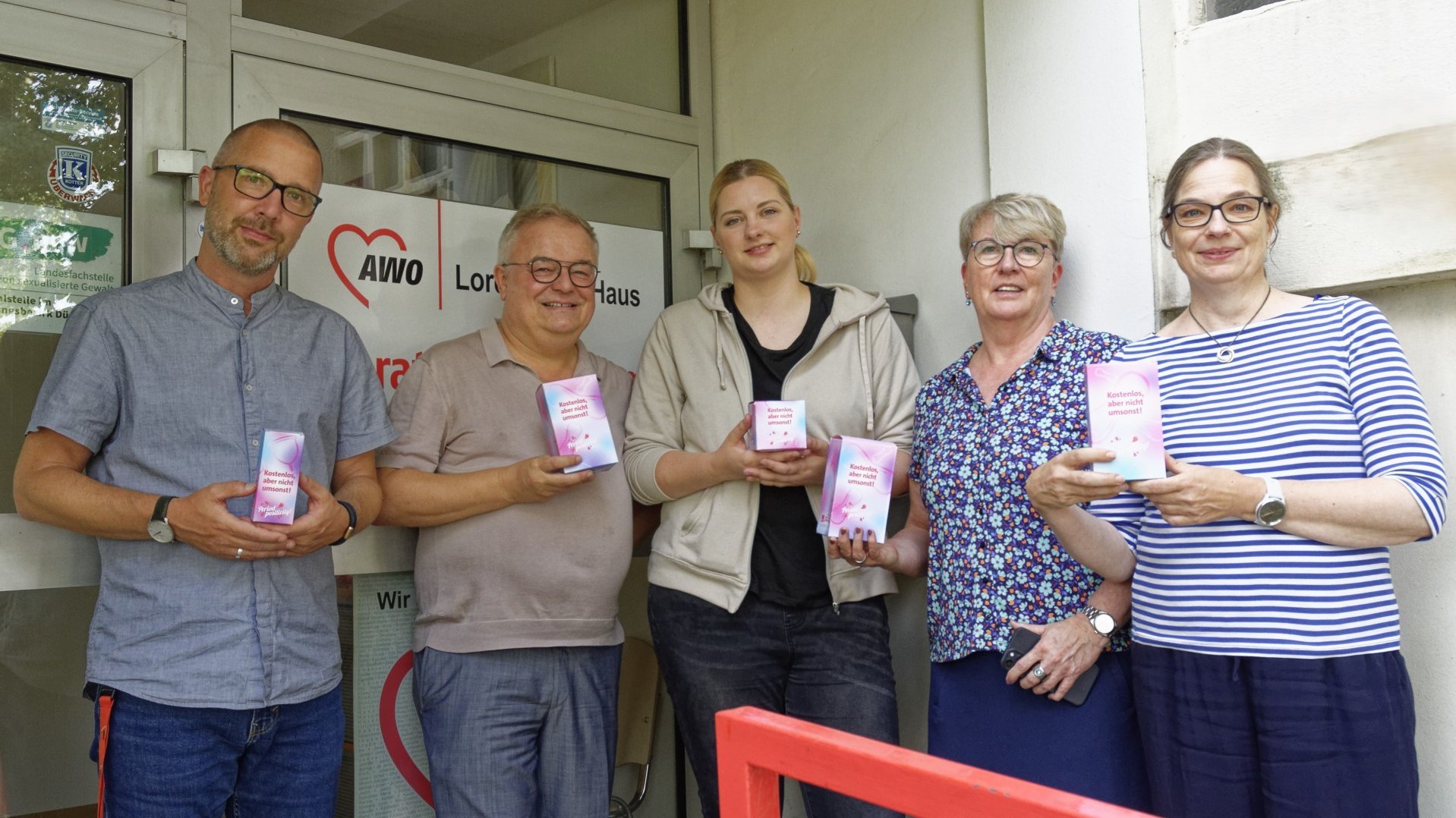 Auf dem Foto sind fünf Personen zu sehen, die vor einem Gebäude stehen. Jede Person hält ein Menstruationsprodukt in der Hand. Neben dem Eingang des Gebäudes ist ein Schild mit dem Logo und dem Namen “AWO” zu sehen, was für “Arbeiterwohlfahrt” steht. Darunter steht “Lore-Agnes-Haus,” was darauf hinweist, dass es sich um eine Einrichtung der AWO namens Lore-Agnes-Haus handelt.