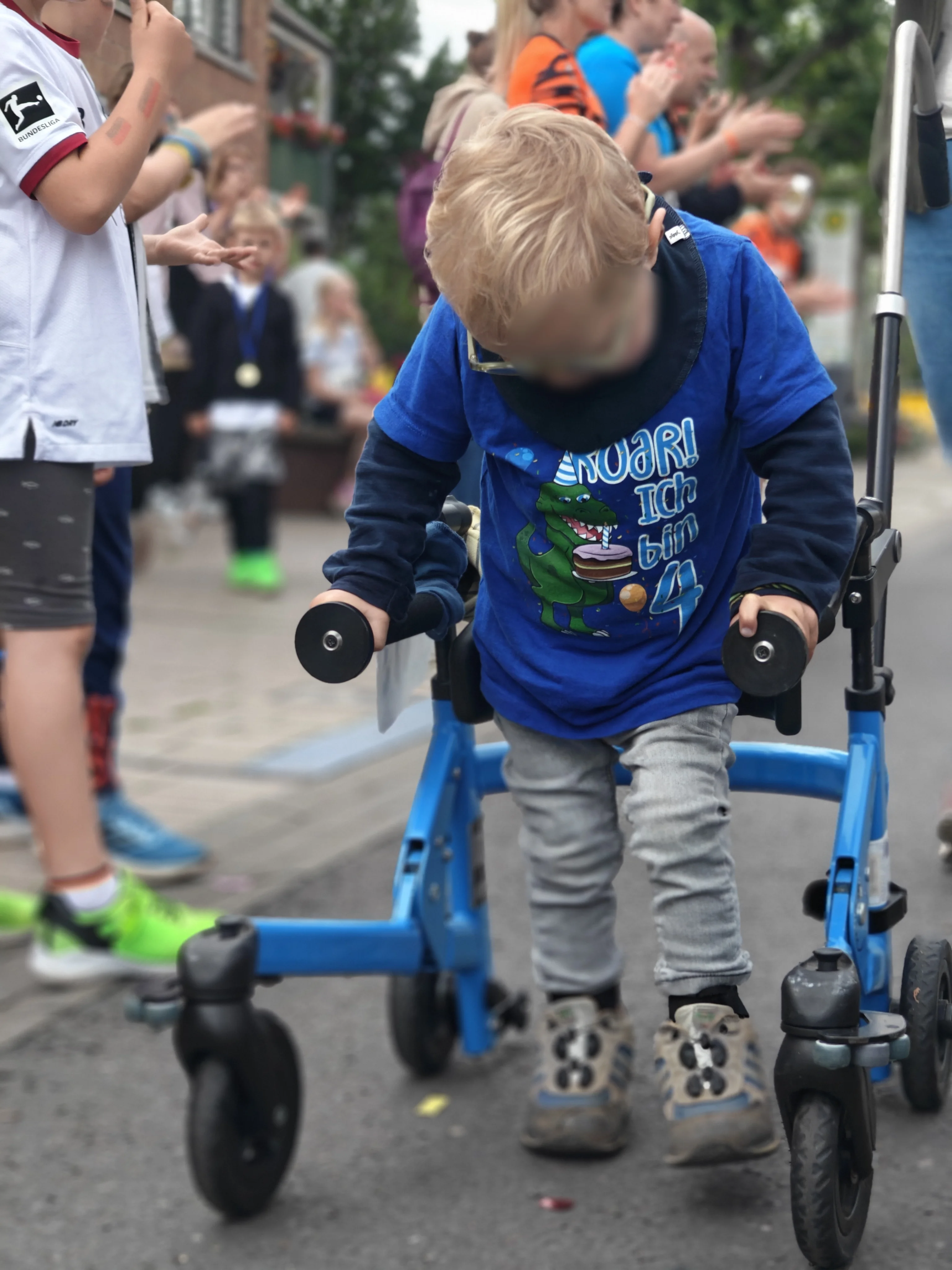 Im Mittelpunkt des Bildes ist ein kleiner Junge zu sehen, der mit einem blauen Gehtrainer auf einer abgesperrten Straße läuft. Er trägt ein blaues T‑Shirt mit einem Dinosaurier und dem Aufdruck „Roar! Ich bin 4“, dazu eine helle Hose und feste Schuhe. Links und im Hintergrund stehen weitere Kinder und Erwachsene am Straßenrand, die offenbar klatschen und anfeuern, was auf eine inklusive Lauf‑ oder Bewegungsaktion hindeutet.