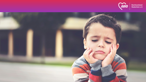 Auf dem Foto ist im Vordergrund ein Kind mit einem bunt gestreiften Pullover zu sehen, dessen Gesicht verpixelt bzw. unkenntlich gemacht ist. ​ Bildinhalt Das Kind stützt den Kopf mit beiden Händen und befindet sich im Freien vor einem unscharfen Hintergrund, der wie ein Gebäude oder eine Hausfassade wirkt. ​ Oben im Bild ist ein breiter Farbverlauf-Balken in Rot‑Pink‑Lila mit dem AWO‑Herz-Logo und der Beschriftung „Bezirksverband Niederrhein e.V.“ zu sehen