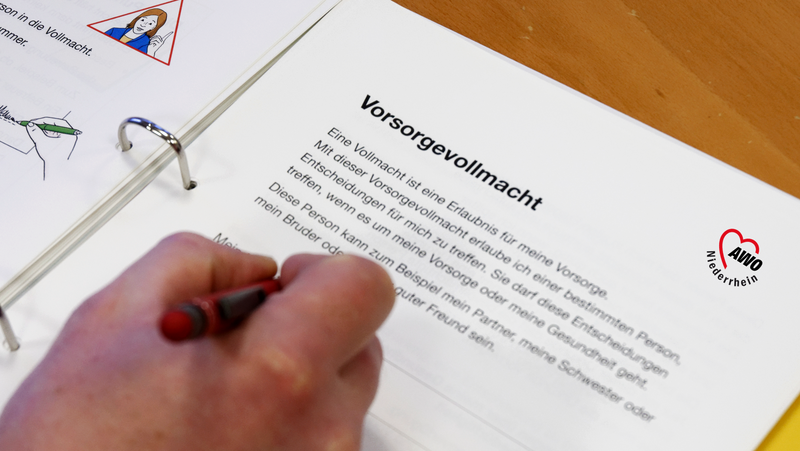 Auf dem Bild ist eine Nahaufnahme des AWO Vorsorge-Ordners Niederrhein zu sehen, bei dem eine Hand mit einem roten Stift gerade etwas in das Ringbuch einträgt. Besonders deutlich erkennbar ist die Titelseite eines Abschnitts mit der Überschrift: Vorsorgevollmacht Darunter steht in leicht verständlicher Sprache die Erklärung der Vorsorgevollmacht: "Eine Vollmacht ist eine Erlaubnis für meine Vorsorge. Mit dieser Vorsorgevollmacht erlaube ich einer bestimmten Person, Entscheidungen für mich zu treffen. Sie darf diese Entscheidungen treffen, wenn es um meine Vorsorge oder meine Gesundheit geht. Diese Person kann zum Beispiel mein Partner, meine Schwester oder mein Bruder oder [unleserlich] guter Freund sein." Rechts auf der Seite ist das Logo des AWO Bezirksverbands Niederrhein in Herzform abgebildet. Im linken oberen Eck sind außerdem schematische Piktogramme aus einem vorhergehenden Abschnitt sichtbar.