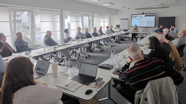 Auf dem Bild ist eine größere Besprechung bzw. Tagung in einem Konferenzraum zu sehen. Mehrere Personen sitzen an Tischen, die U‑förmig gestellt sind, und hören einer Präsentation zu. Vorne im Raum läuft auf einem großen Bildschirm eine Präsentation, daneben stehen Flipchart und Pinnwand. Auf den Tischen liegen Laptops, Notizblöcke, Gläser und Wasserkarafen