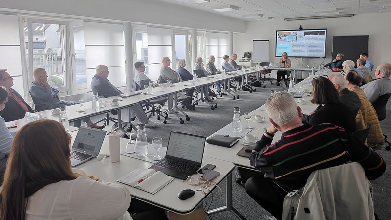 Auf dem Bild ist eine größere Besprechung bzw. Tagung in einem Konferenzraum zu sehen. Mehrere Personen sitzen an Tischen, die U‑förmig gestellt sind, und hören einer Präsentation zu. Vorne im Raum läuft auf einem großen Bildschirm eine Präsentation, daneben stehen Flipchart und Pinnwand. Auf den Tischen liegen Laptops, Notizblöcke, Gläser und Wasserkarafen