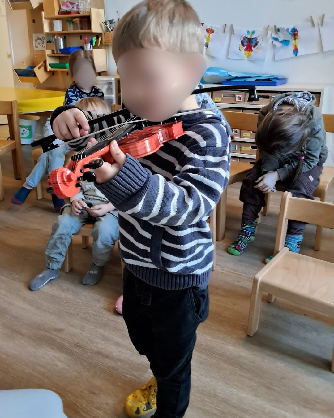 Auf dem Bild ist ein kleiner Junge (Kleinkind/Kindergartenalter) zu sehen, der eine rote Geige spielt. Er trägt einen blau-weiß gestreiften Pullover und dunkle Hose. Im Hintergrund sind weitere Kinder sowie typisches Kindergarten-Mobiliar (Holzstühle, Regale) und an der Wand hängende Kinderzeichnungen zu erkennen.
