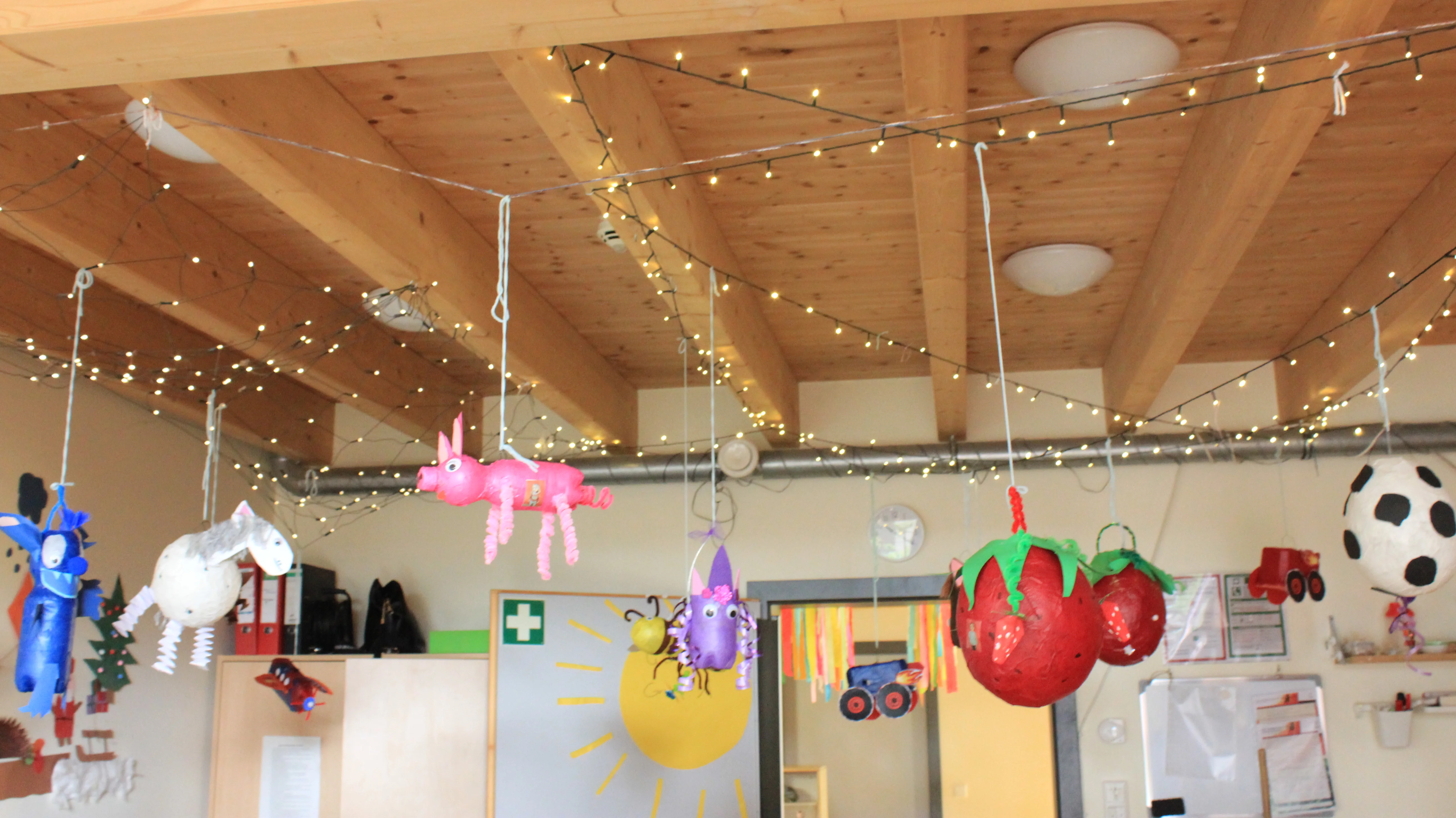 Zu sehen ist ein Gruppenraum in einer Kita oder Grundschule. An einer Holzdecke mit sichtbaren Balken hängen zahlreiche selbst gebastelte Figuren und Laternen: unter anderem ein pinkes Tier, ein Einhorn, eine große Erdbeere, ein Fußball sowie weitere bunte Fantasiefiguren. Zusätzlich sind Lichterketten gespannt, im Hintergrund erkennt man Schränke, eine Türgestaltung mit Sonne und Regenbogenbändern sowie einen Erste-Hilfe-Hinweis. Die Szene wirkt wie eine herbstliche Bastel- oder Laternenausstellung in einer pädagogischen Einrichtung.
