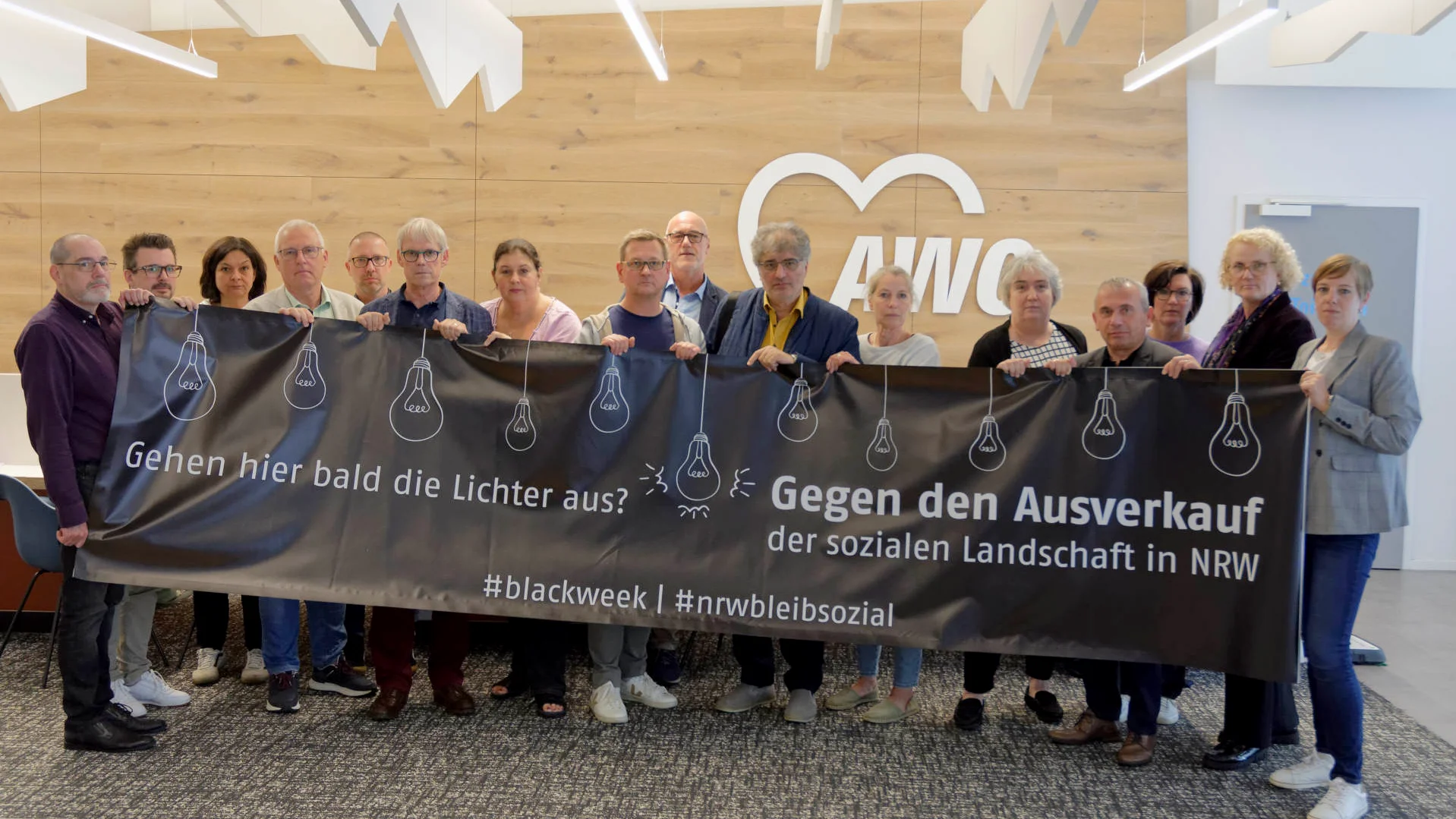 Auf dem Foto ist eine Gruppe von Personen zu sehen, die drinnen stehen und ein großes Banner halten. Auf dem Banner steht Text in deutscher Sprache und einige grafische Elemente. Der Text auf dem Banner lautet: “Gehen hier bald die Lichter aus? Gegen den Ausverkauf der sozialen Landschaft in NRW #blackweek #niewiederbilligsozial”. Dies übersetzt sich mit “Will the lights go out here soon? Against the sell-out of the social landscape in NRW (North Rhine-Westphalia) #blackweek #neveragaincheapsocial”. Der Banner enthält Grafiken von Glühbirnen mit einem Kreuz durch sie, was auf etwas mit dem Ausschalten von Licht oder dem Verlust von Licht hinweisen könnte. Dies könnte metaphorisch für das Bewusstsein oder die Aufmerksamkeit für ein Problem stehen. Der Hashtag deutet darauf hin, dass dies Teil einer Kampagne oder Bewegung im Zusammenhang mit sozialen Themen in Nordrhein-Westfalen, Deutschland, ist.