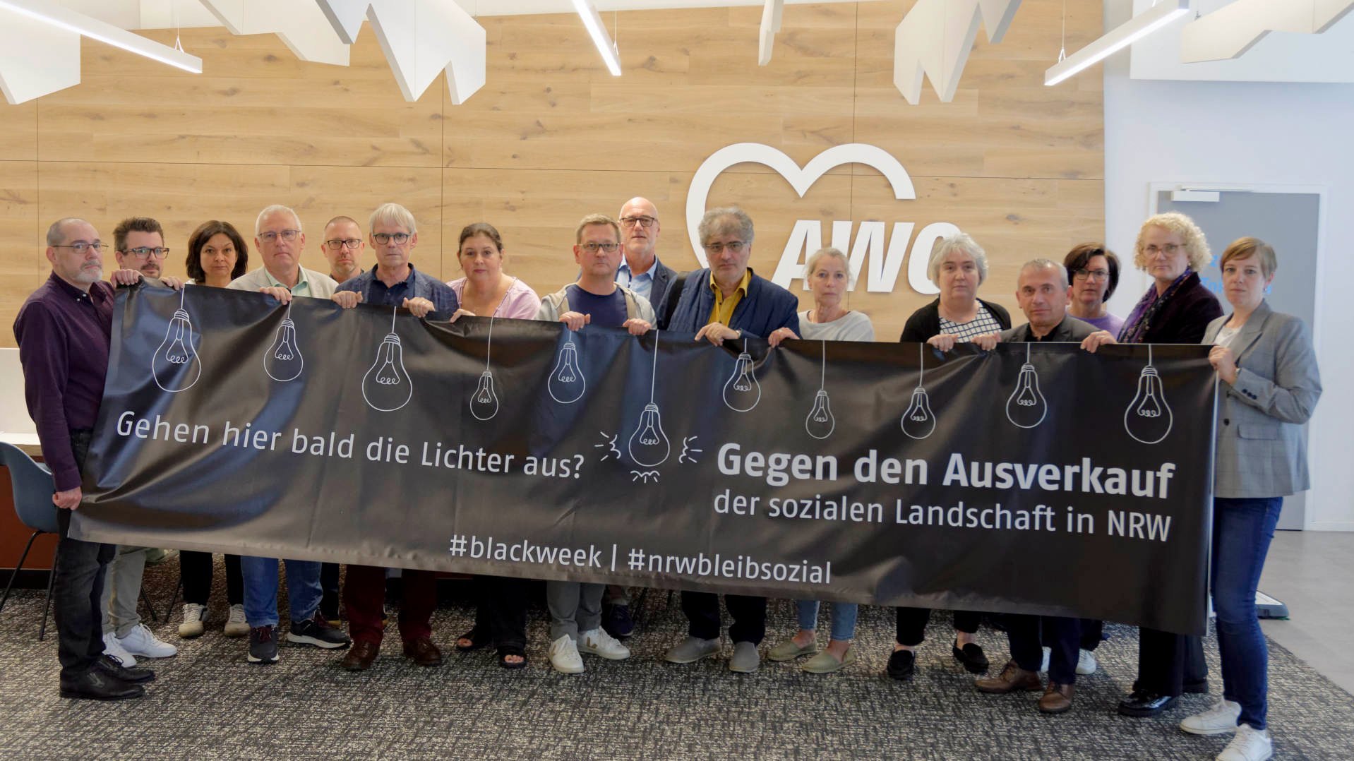 Auf dem Foto ist eine Gruppe von Personen zu sehen, die drinnen stehen und ein großes Banner halten. Auf dem Banner steht Text in deutscher Sprache und einige grafische Elemente. Der Text auf dem Banner lautet: “Gehen hier bald die Lichter aus? Gegen den Ausverkauf der sozialen Landschaft in NRW #blackweek #niewiederbilligsozial”. Dies übersetzt sich mit “Will the lights go out here soon? Against the sell-out of the social landscape in NRW (North Rhine-Westphalia) #blackweek #neveragaincheapsocial”. Der Banner enthält Grafiken von Glühbirnen mit einem Kreuz durch sie, was auf etwas mit dem Ausschalten von Licht oder dem Verlust von Licht hinweisen könnte. Dies könnte metaphorisch für das Bewusstsein oder die Aufmerksamkeit für ein Problem stehen. Der Hashtag deutet darauf hin, dass dies Teil einer Kampagne oder Bewegung im Zusammenhang mit sozialen Themen in Nordrhein-Westfalen, Deutschland, ist. 