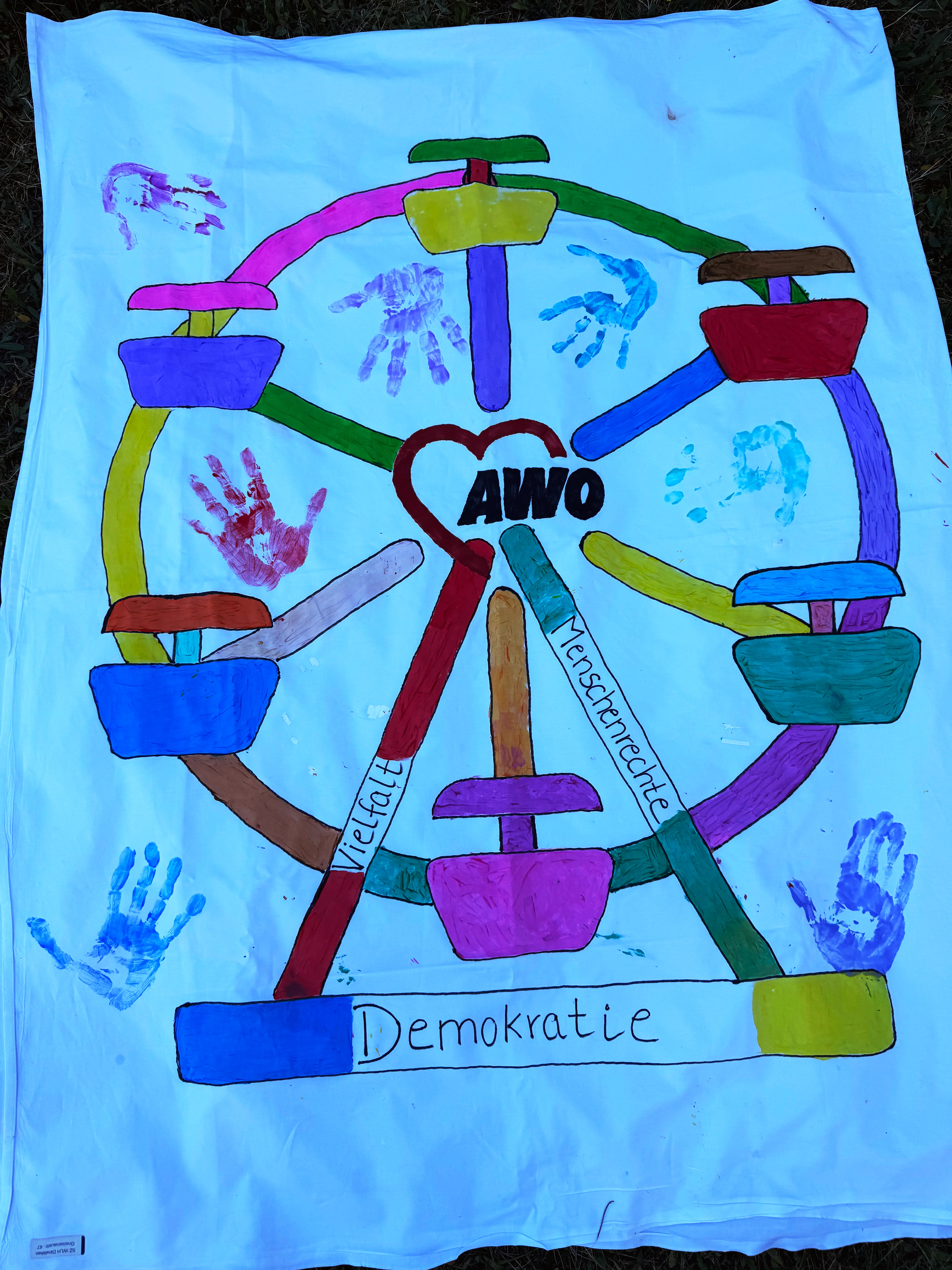 Auf dem Bild ist ein bunt gemaltes Riesenrad zu sehen, das als Symbol für Demokratie, Vielfalt und Menschenrechte gestaltet ist. In der Mitte des Riesenrads steht ein rotes Herz mit der Aufschrift „AWO“, das auf die Arbeiterwohlfahrt verweist. Die Streben des Riesenrads sind in verschiedenen Farben gehalten; auf zwei der Streben stehen die Wörter „Vielfalt“ und „Menschenrechte“, unten quer über den Unterbau ist ein Balken mit der Aufschrift „Demokratie“ angebracht. Rund um das Riesenrad und dazwischen sind mehrere farbige Handabdrücke (rot, blau, türkis, violett usw.) zu sehen, die darauf hindeuten, dass Kinder oder mehrere Personen gemeinsam daran gearbeitet haben. Der Hintergrund ist ein weißes Tuch oder Banner, das offenbar draußen auf dem Boden ausgelegt wurde.