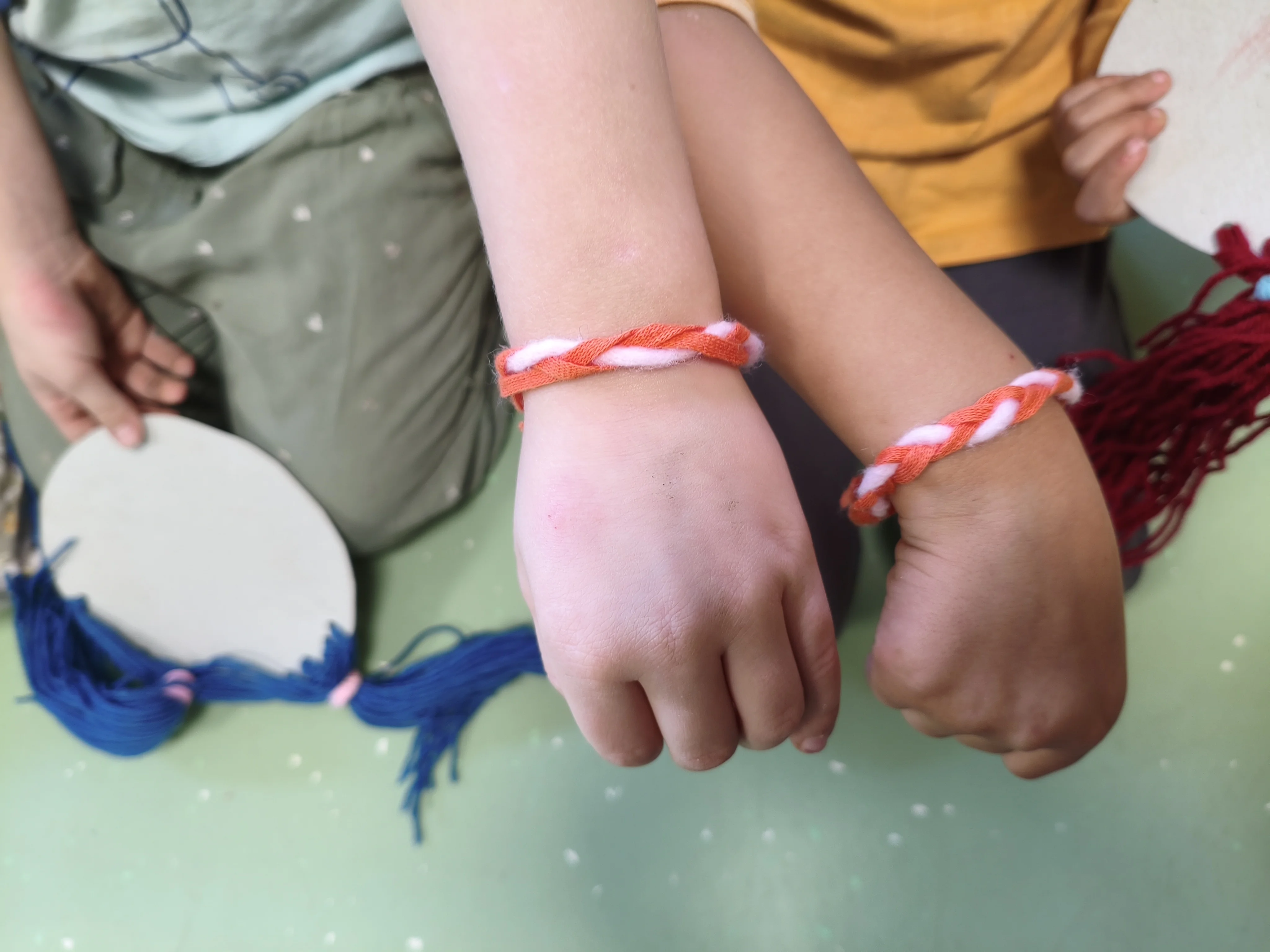 Auf dem Bild zeigen zwei Kinder ihre selbst gemachten Armbänder. Die Bänder bestehen aus miteinander verdrehten orangefarbenen und hellrosa Strängen und werden an den Handgelenken nebeneinander präsentiert. Die Gesichter der Kinder sind nicht zu sehen. Daneben halten sie helle, rundliche Bastelvorlagen aus Pappe, an denen blaue beziehungsweise dunkelrote Wollfäden befestigt sind. Im Hintergrund sieht man einen grünen, hell gesprenkelten Boden.