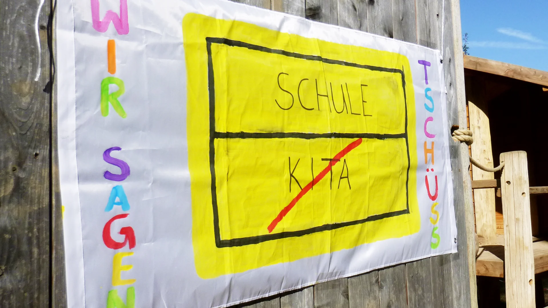 Das Foto zeigt ein handgemachtes Schild, das an einer Holzoberfläche befestigt ist. Das Schild hat einen weißen Hintergrund mit farbenfroher Beschriftung und einem gelb umrandeten Rechteck in der Mitte. Im Rechteck steht oben das Wort “SCHULE” und unten “KITA”, beide in schwarzen Buchstaben. Über dem Rechteck steht “WIR SAGEN” in lila Buchstaben, und rechts davon steht “TSCHÜSS” in grünen Buchstaben.