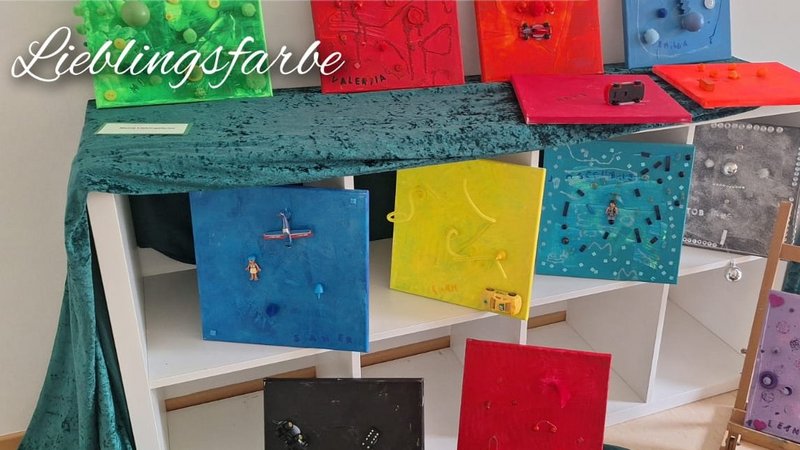 Auf dem Bild ist eine kleine Ausstellung bzw. Vernissage mit Kinder‑Kunstwerken zu sehen. Lieblingsfarbe Oben sieht man mehrere quadratische Leinwände in unterschiedlichen, sehr kräftigen Farben (Grün, Rot, Blau, Gelb, Orange, Schwarz, Türkis, Lila etc.), die auf einem Regal und davor präsentiert werden. Viele sind mit kleinen Objekten, Strukturen und Reliefs beklebt oder bemalt, passend zum Schriftzug „Lieblingsfarbe“ im Bild. Lieblingstier Links unten ist ein Bereich mit goldfarbenen Ton‑ oder Gipsfiguren und ‑köpfen, die auf kleinen Holzsockeln stehen und auf einem braunen Stoff arrangiert sind. Daneben steht der Schriftzug „Lieblingstier“, was darauf hindeutet, dass es sich um Skulpturen von Lieblingstieren der Kinder handelt. Selbstporträt Rechts unten sieht man mehrere bunte Bilder von Gesichtern, typische Kinder‑Selbstporträts mit großen Augen, Mund und Haaren, teils mit gemustertem Hintergrund. Diese liegen oder stehen auf braunem Stoff, und im Bild ist der Schriftzug „Selbstporträt“ eingeblendet, außerdem sind einige kleine Schildchen zu erkennen, vermutlich mit Namen oder Werktiteln der Kinder.