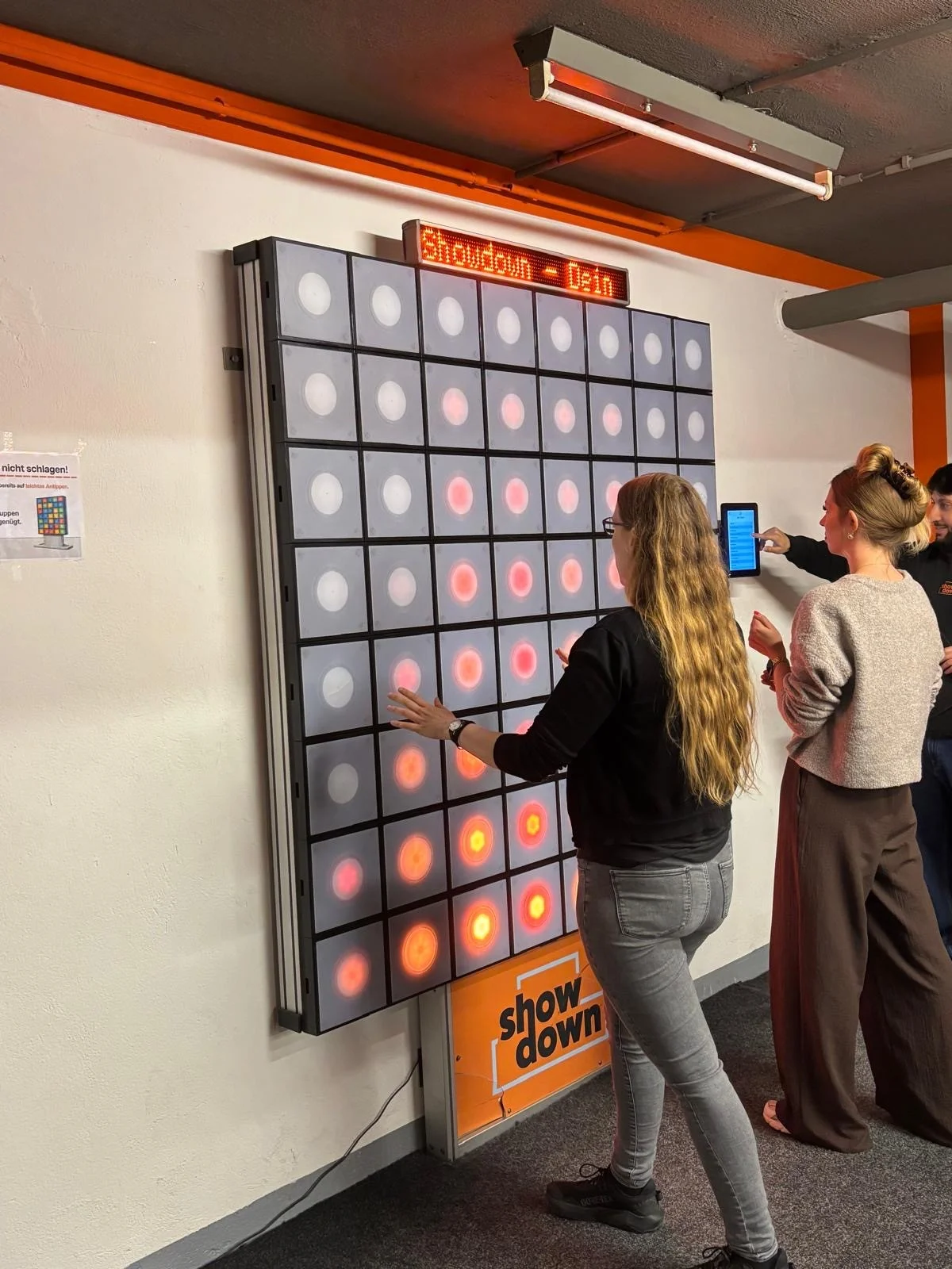 Auf dem Foto sind mehrere Personen in einem Raum zu sehen, die ein großes, senkrecht an der Wand montiertes Reaktions‑ oder Lichtspiel bedienen. Die vordere Person tippt mit den Händen auf leuchtende Felder einer Matrix aus Lichtplatten, während im Hintergrund weitere Personen danebenstehen und offenbar das Spiel oder die Anzeige bedienen.