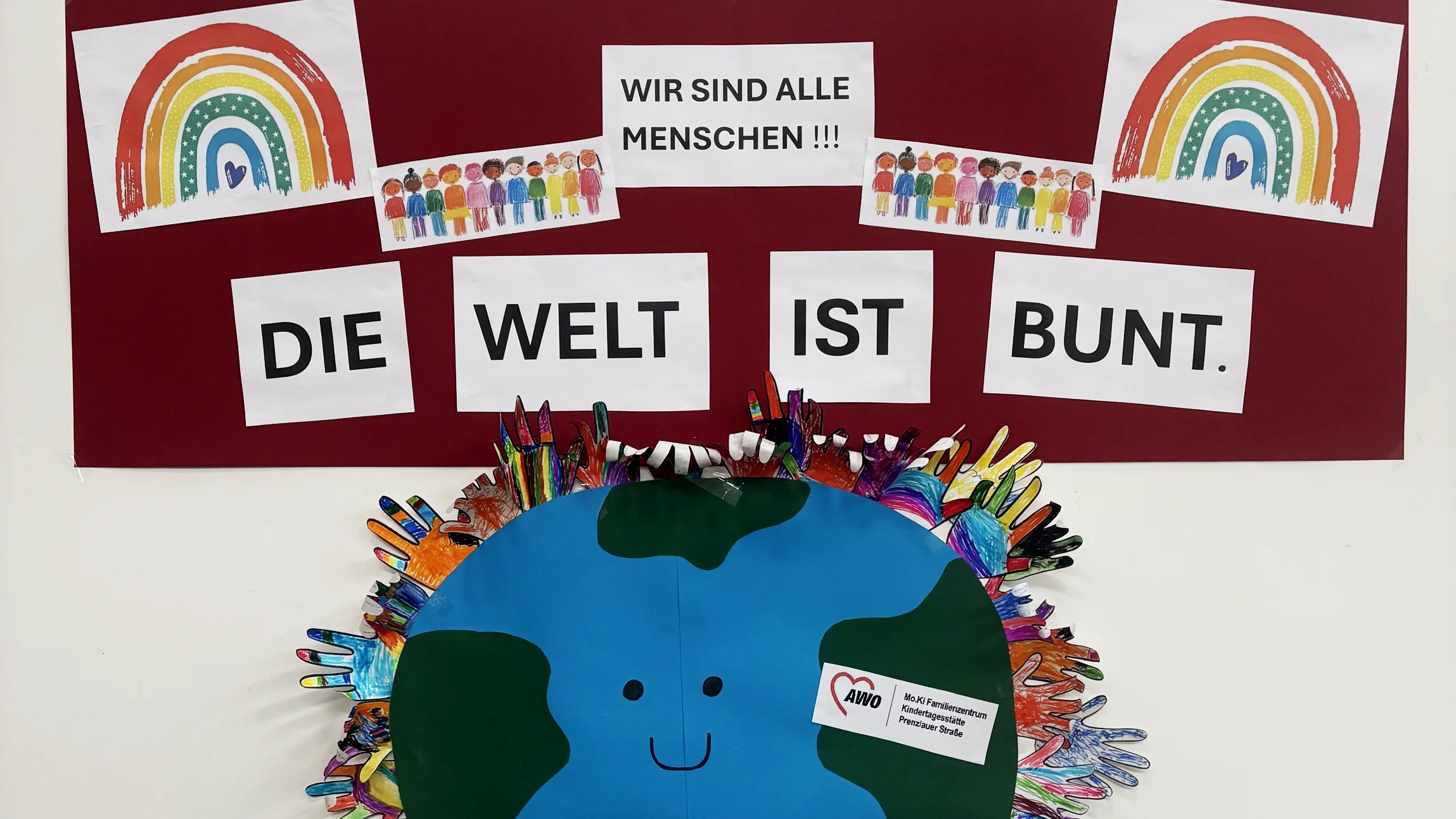 Auf dem Foto ist eine kreative Bastel- und Kunstarbeit zum Thema Vielfalt und Toleranz zu sehen, die von Kindern gestaltet wurde: Plakat (dunkelroter Hintergrund) mit den Aufschriften: „WIR SIND ALLE MENSCHEN!!!" „DIE WELT IST BUNT." Zwei Regenbögen (links und rechts), die von Kindern gemalt wurden. Kleine Bildkarten mit gezeichneten, bunten Menschenfiguren in unterschiedlichen Hautfarben – als Symbol für Diversität. Eine große, runde Weltkugel aus Papier/Pappe in Blau und Grün mit einem aufgemalten Smiley-Gesicht – umrahmt von vielen bunt bemalten Handabdrücken der Kinder in allen möglichen Farben.