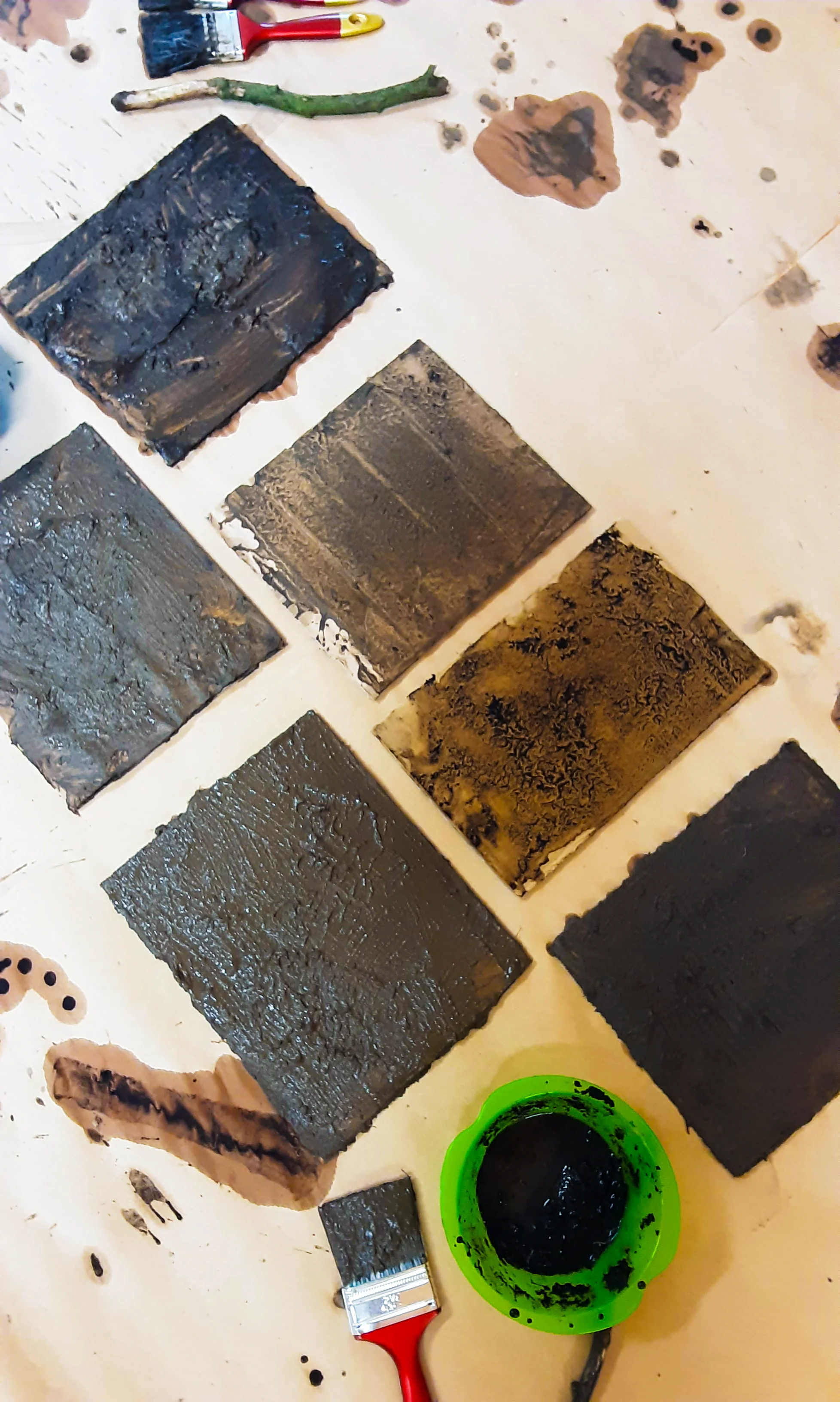 Auf dem Bild sind mehrere rechteckige Platten oder Papierstücke zu sehen, die mit dunkler Erde bestrichen wurden. Daneben liegen Pinsel, ein grüner Zweig und ein Schälchen mit schwarzer, breiiger Farbe. Auf der Unterlage befinden sich außerdem verschiedene Farb- und Erdspuren sowie einfache Abdrücke.