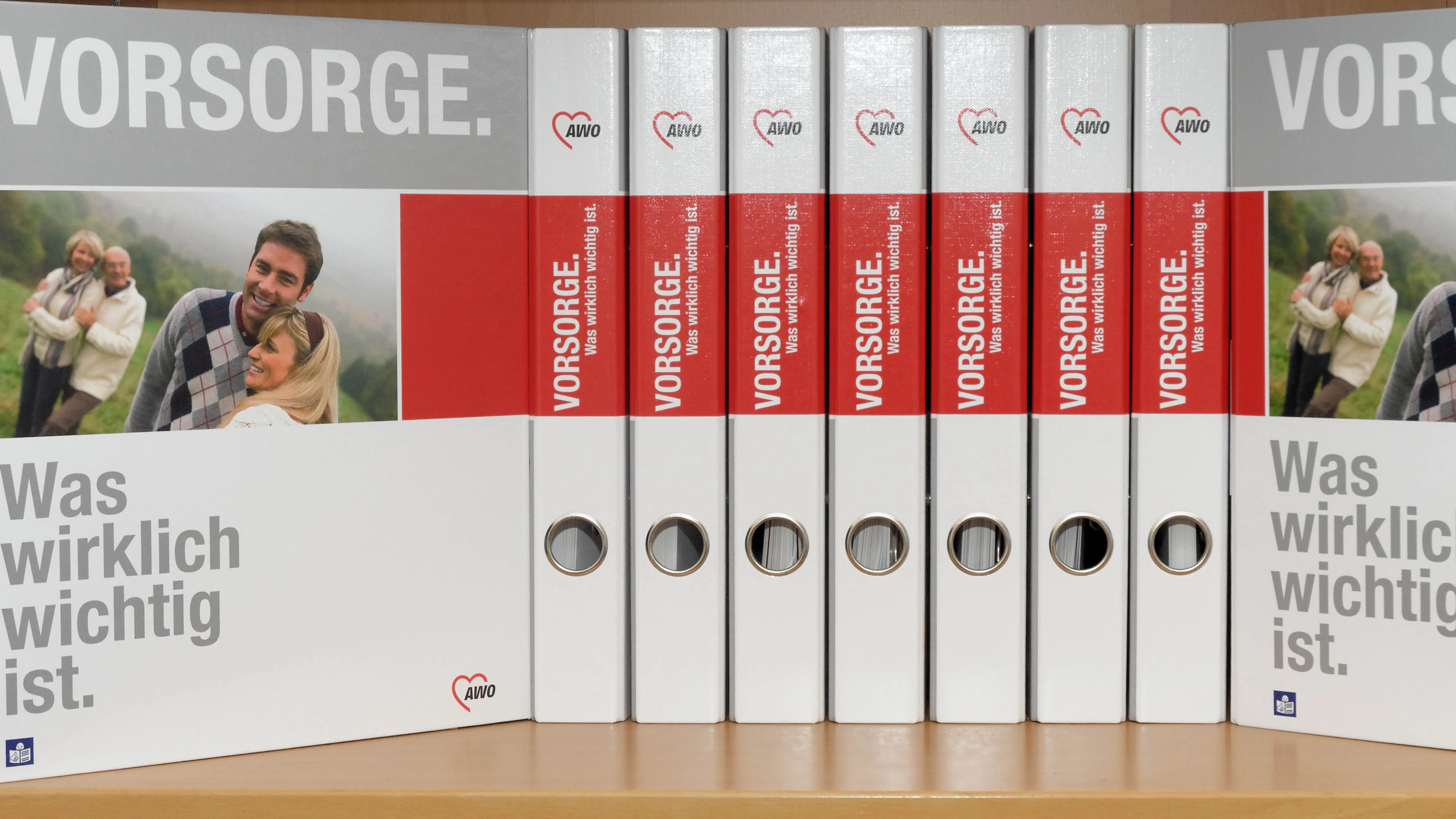 Auf dem Foto sind mehrere weiße Ordner (Aktenordner) in einem Regal zu sehen. Die Ordner sind einheitlich gestaltet und tragen alle den Aufdruck: Oben groß in weiß auf grauem Hintergrund: „VORSORGE.“ Seitlich unten: „Was wirklich wichtig ist.“ Mit einem Logo der AWO (Arbeiterwohlfahrt), einer deutschen Wohlfahrtsorganisation. Auf den Ordnerrücken steht ebenfalls „VORSORGE.“ sowie weitere kleinere Beschriftung, die wahrscheinlich auf Inhalte oder Kapitel hinweist. Zudem ist auf den äußeren Ordnern jeweils ein Bild eines Paares oder älterer Menschen zu sehen, was auf das Thema „Vorsorge“ im Sinne von Altersvorsorge, Patientenverfügung, Pflege oder ähnlichem hinweist.