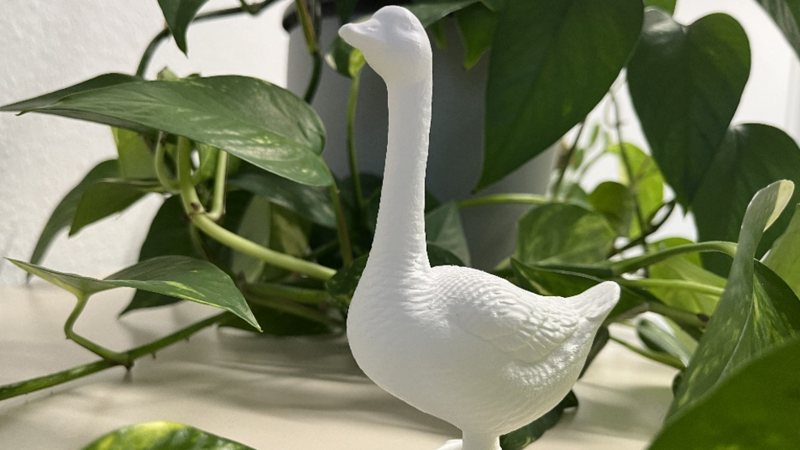 Auf dem Bild steht eine weiße Gänsefigur zwischen üppigen grünen Zimmerpflanzen auf einer hellen Fensterbank. Sie hat einen langen, aufrechten Hals, einen leicht nach links gerichteten Kopf und eine strukturierte Oberfläche, die Federn nachbildet.