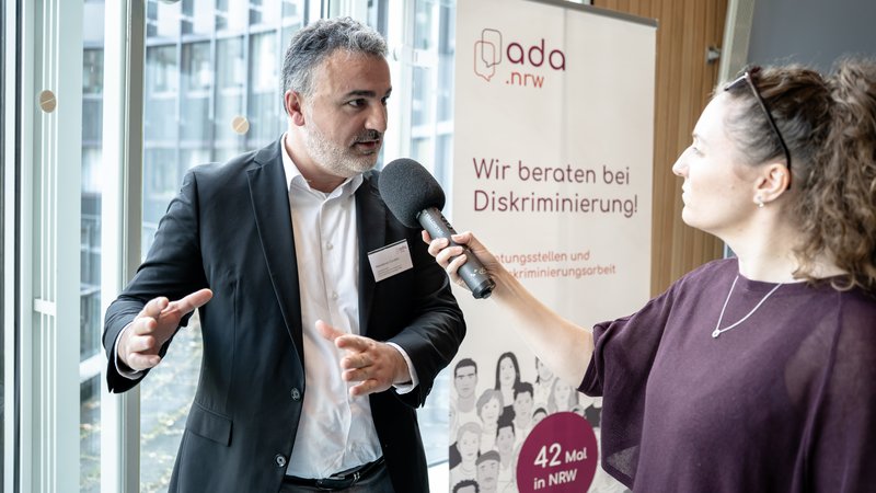 Das Foto zeigt eine Interviewsituation bei einer Veranstaltung von ada.nrw. Ein Mann im Anzug spricht in ein Handmikrofon, das ihm eine Frau entgegenhält. Im Hintergrund ist ein Roll-up-Banner zu sehen. Darauf steht: „Wir beraten bei Diskriminierung!“ sowie der Hinweis auf „42 Mal in NRW“. Die Szene spielt in einem modernen, hellen Gebäude mit großen Fensterfronten