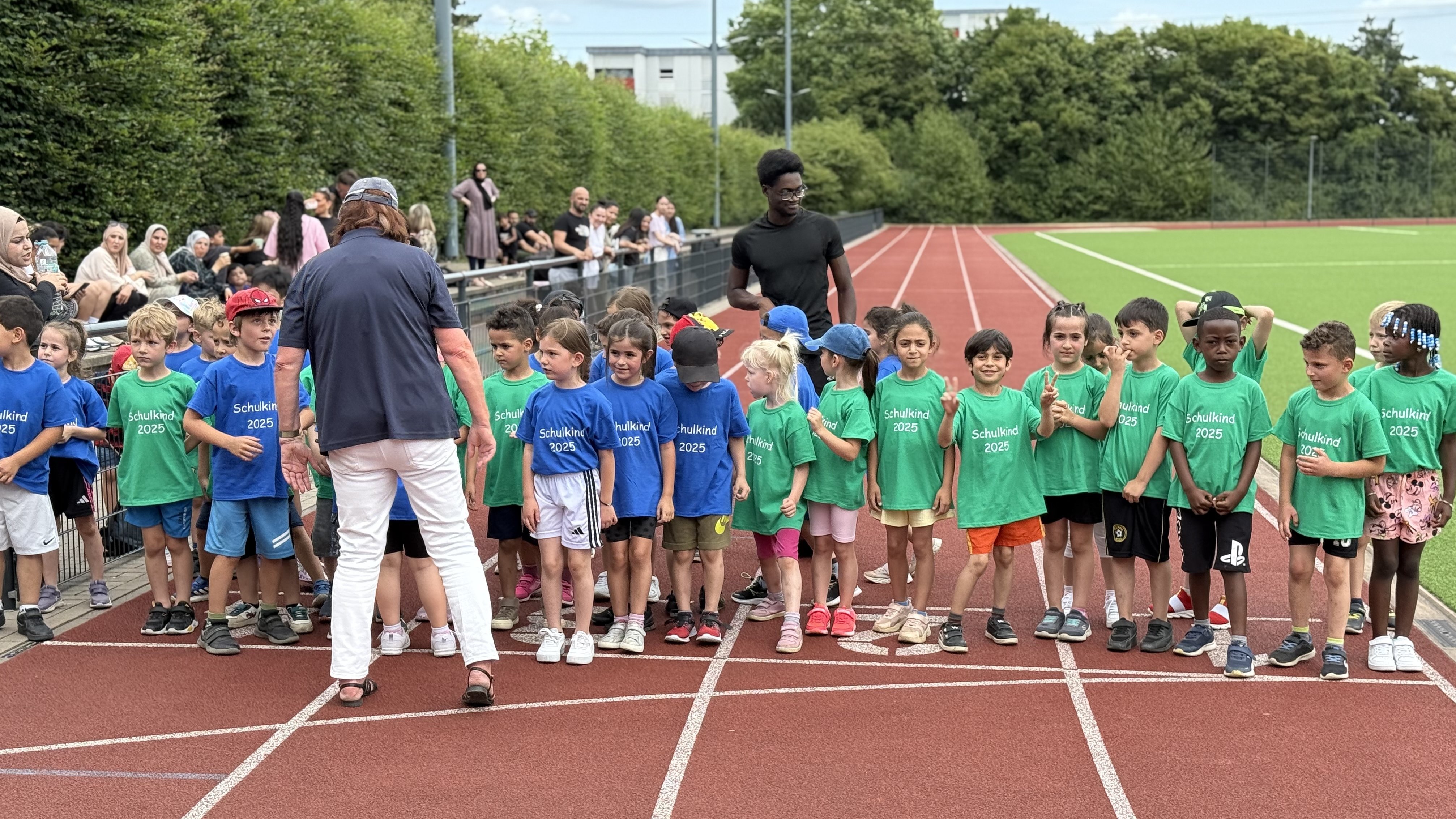 Auf dem Foto ist eine Gruppe von Kindern auf einer Laufbahn zu sehen, die in zwei Reihen aufgestellt sind. Die Kinder tragen entweder blaue oder grüne T-Shirts. Im Hintergrund stehen Erwachsene, die den Kindern zugewandt sind, und es scheint sich um eine Sportveranstaltung zu handeln, möglicherweise einen Lauf oder Wettkampf. Die Umgebung deutet auf ein Sportstadion mit einer Tartanbahn und Grünflächen hin.