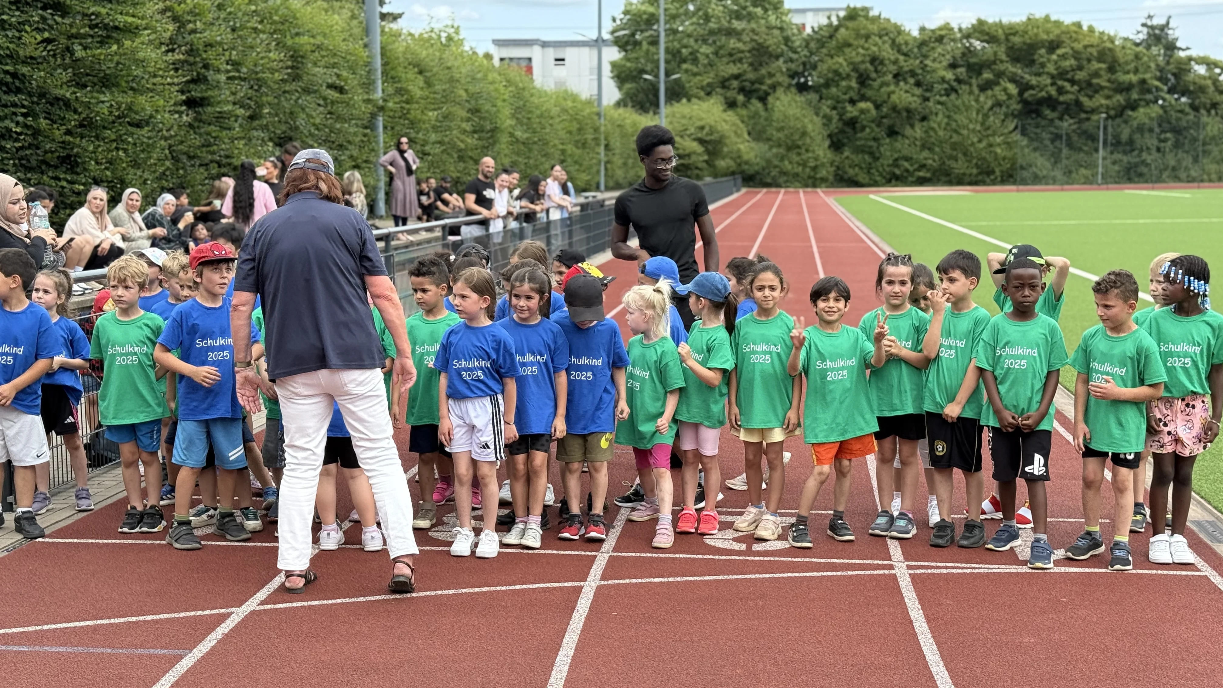 Auf dem Foto ist eine Gruppe von Kindern auf einer Laufbahn zu sehen, die in zwei Reihen aufgestellt sind. Die Kinder tragen entweder blaue oder grüne T-Shirts. Im Hintergrund stehen Erwachsene, die den Kindern zugewandt sind, und es scheint sich um eine Sportveranstaltung zu handeln, möglicherweise einen Lauf oder Wettkampf. Die Umgebung deutet auf ein Sportstadion mit einer Tartanbahn und Grünflächen hin.