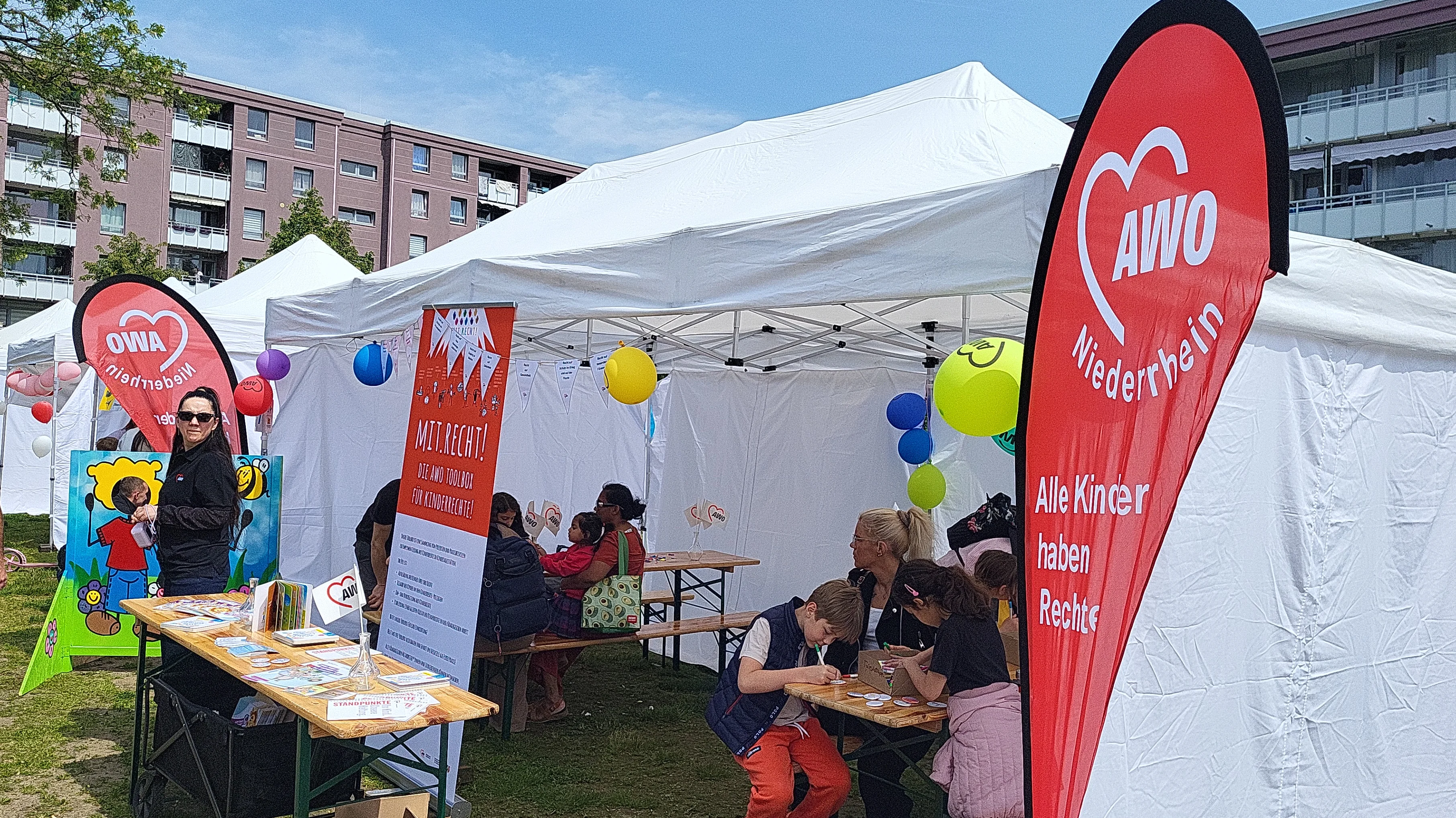 Auf dem Bild ist ein AWO-Stand bei einer Outdoor-Veranstaltung zu sehen, vermutlich einem Kinder- oder Familienfest. Vor mehreren weißen Pavillons stehen Tische mit Infomaterial, es gibt bunte Luftballons, AWO‑Beachflags mit dem Hinweis „Alle Kinder haben Rechte“, ein Kinder-Mitmachaufsteller mit zwei Comicfiguren sowie Sitzbänke, an denen Kinder gemeinsam mit Erwachsenen malen und basteln.