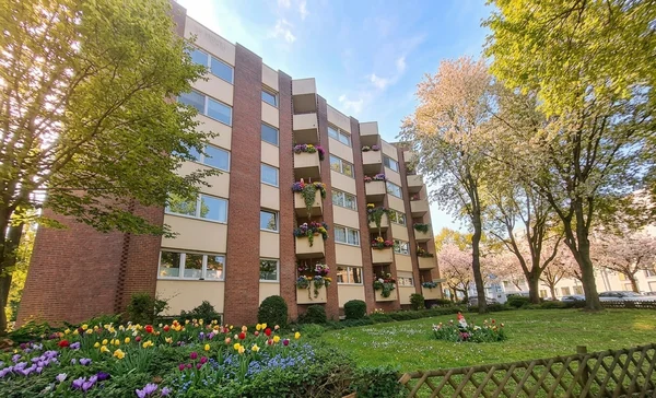 Auf dem Foto ist ein mehrstöckiges Wohnhaus zu sehen, mit Balkonen an der Vorderseite. Davor liegt eine größere Rasenfläche mit Büschen und mehreren hohen Bäumen, eingefasst von einem niedrigen Holzzaun entlang des Gehwegs.