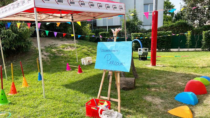 Auf dem Foto ist ein bunter Geschicklichkeits-Parcours im Freien zu sehen, vermutlich bei einem Sommerfest der AWO (Arbeiterwohlfahrt), erkennbar am rot-weißen Pavillon mit AWO-Logo. Der Parcours befindet sich auf einer Rasenfläche und besteht aus verschiedenen Spiel- und Bewegungselementen: Ein handgemaltes Schild auf einer Staffelei mit der Aufschrift „Geschicklichkeits-Parcours“, Bunte Hütchen mit Stangen, Kunststoff-Elemente zum Balancieren (in Rot, Blau, Orange, Gelb und Grün), Hula-Hoop-Reifen, Bunte Wimpelketten zur Dekoration, Rote Kisten mit Materialien und ein Netzbeutel, Im Hintergrund ist ein Mehrfamilienhaus und ein sandiger Hügel zu sehen. Die Szene vermittelt eine fröhliche, kindgerechte Atmosphäre – ideal für Spiel, Bewegung und Spaß im Rahmen einer Familien- oder Kita-Veranstaltung.