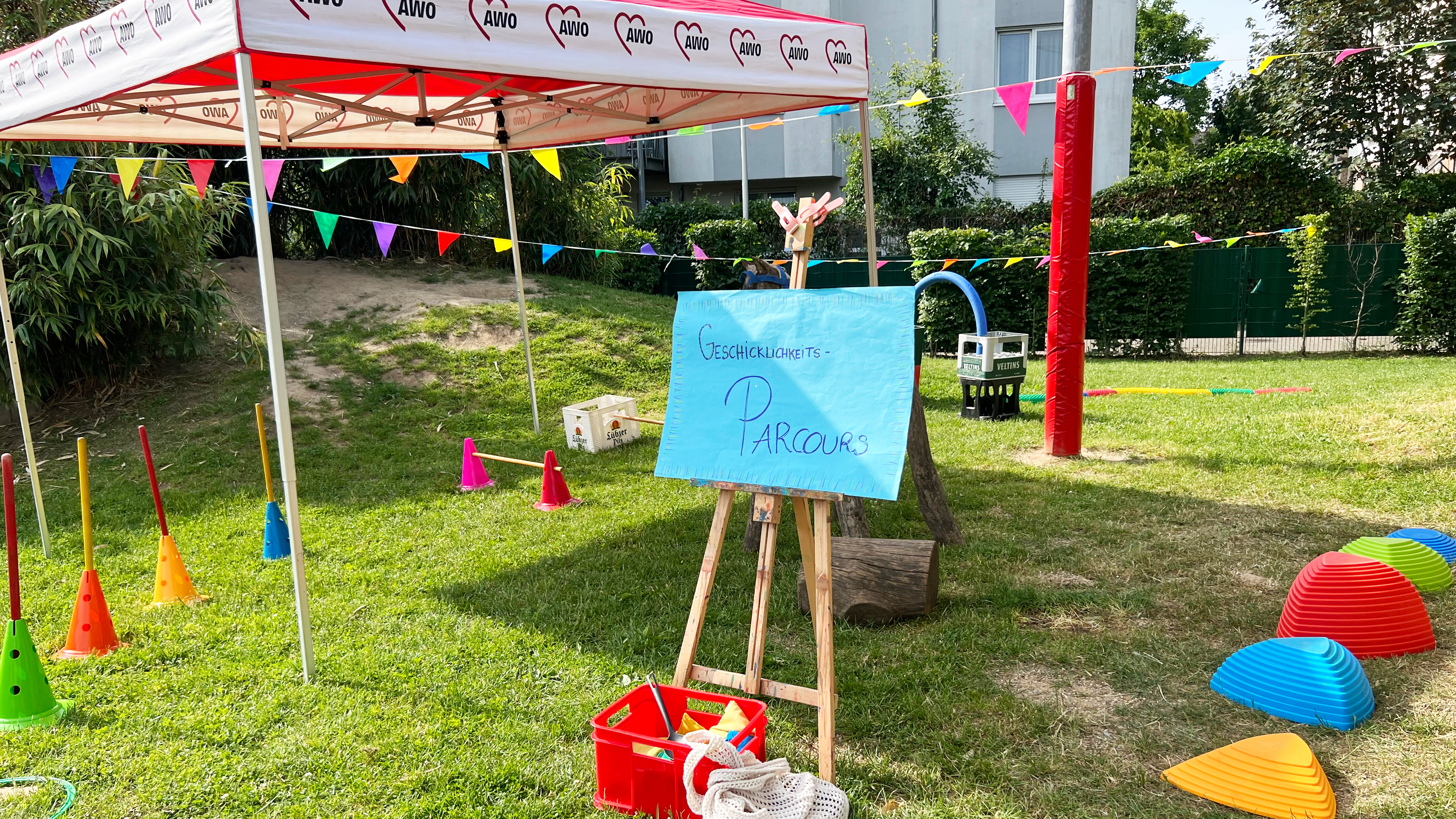 Auf dem Foto ist ein bunter Geschicklichkeits-Parcours im Freien zu sehen, vermutlich bei einem Sommerfest der AWO (Arbeiterwohlfahrt), erkennbar am rot-weißen Pavillon mit AWO-Logo. Der Parcours befindet sich auf einer Rasenfläche und besteht aus verschiedenen Spiel- und Bewegungselementen: Ein handgemaltes Schild auf einer Staffelei mit der Aufschrift „Geschicklichkeits-Parcours“, Bunte Hütchen mit Stangen, Kunststoff-Elemente zum Balancieren (in Rot, Blau, Orange, Gelb und Grün), Hula-Hoop-Reifen, Bunte Wimpelketten zur Dekoration, Rote Kisten mit Materialien und ein Netzbeutel, Im Hintergrund ist ein Mehrfamilienhaus und ein sandiger Hügel zu sehen. Die Szene vermittelt eine fröhliche, kindgerechte Atmosphäre – ideal für Spiel, Bewegung und Spaß im Rahmen einer Familien- oder Kita-Veranstaltung.