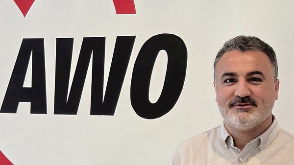 Auf dem Foto ist ein Mann zu sehen, der vor einer weißen Wand mit einem großen, roten und schwarzen Logo steht. Das Logo zeigt ein Herz und die Buchstaben AWO. Der Mann trägt ein helles, beiges oder graues Hemd, hat einen Bart und schaut in die Kamera.