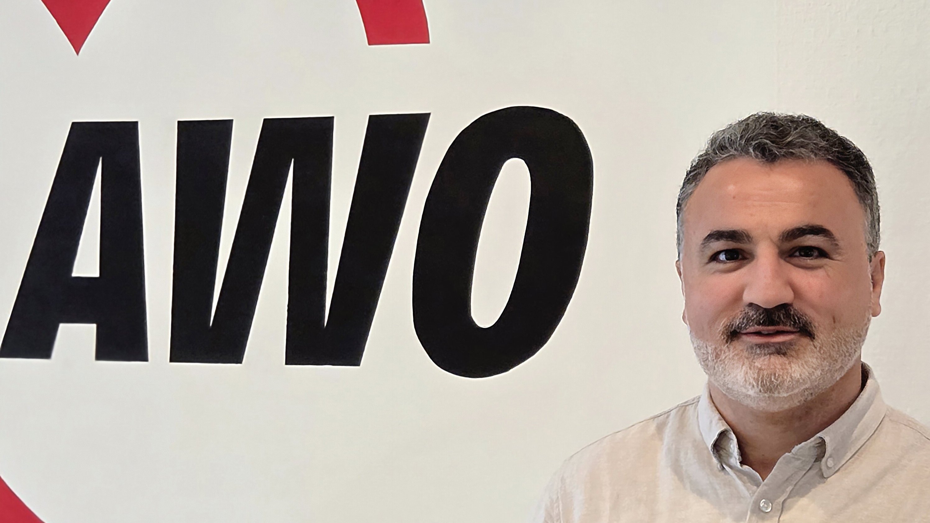  Auf dem Foto ist ein Mann zu sehen, der vor einer weißen Wand mit einem großen, roten und schwarzen Logo steht. Das Logo zeigt ein Herz und die Buchstaben AWO. Der Mann trägt ein helles, beiges oder graues Hemd, hat einen Bart und schaut in die Kamera.
