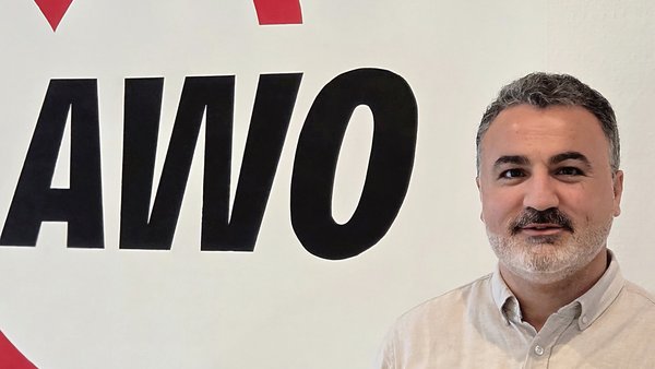  Auf dem Foto ist ein Mann zu sehen, der vor einer weißen Wand mit einem großen, roten und schwarzen Logo steht. Das Logo zeigt ein Herz und die Buchstaben AWO. Der Mann trägt ein helles, beiges oder graues Hemd, hat einen Bart und schaut in die Kamera.