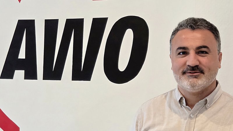  Auf dem Foto ist ein Mann zu sehen, der vor einer weißen Wand mit einem großen, roten und schwarzen Logo steht. Das Logo zeigt ein Herz und die Buchstaben AWO. Der Mann trägt ein helles, beiges oder graues Hemd, hat einen Bart und schaut in die Kamera.
