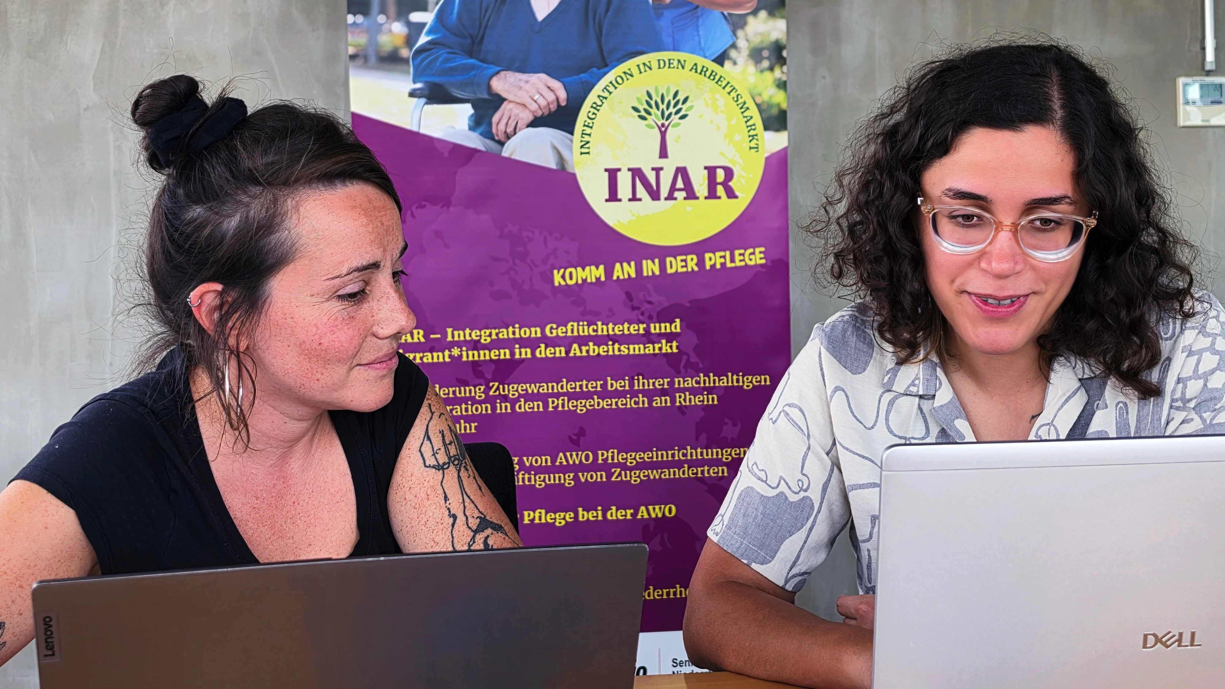 Auf dem Bild sind zwei Frauen zu sehen, die nebeneinander an einem Tisch mit aufgeklappten Laptops sitzen. Die Frau links blickt zur Frau rechts, die auf ihren Bildschirm schaut. Die Szene wirkt wie eine gemeinsame Arbeits- oder Beratungssituation. Im Hintergrund steht ein großes Banner des Projekts „INAR – Integration in den Arbeitsmarkt“ mit dem Slogan „Komm an in der Pflege“. Darauf ist eine Pflegekraft mit einem älteren Mann im Rollstuhl abgebildet. Der weitere Bannertext thematisiert die Integration von Geflüchteten und Migrant*innen in den Arbeitsmarkt beziehungsweise in den Pflegebereich.