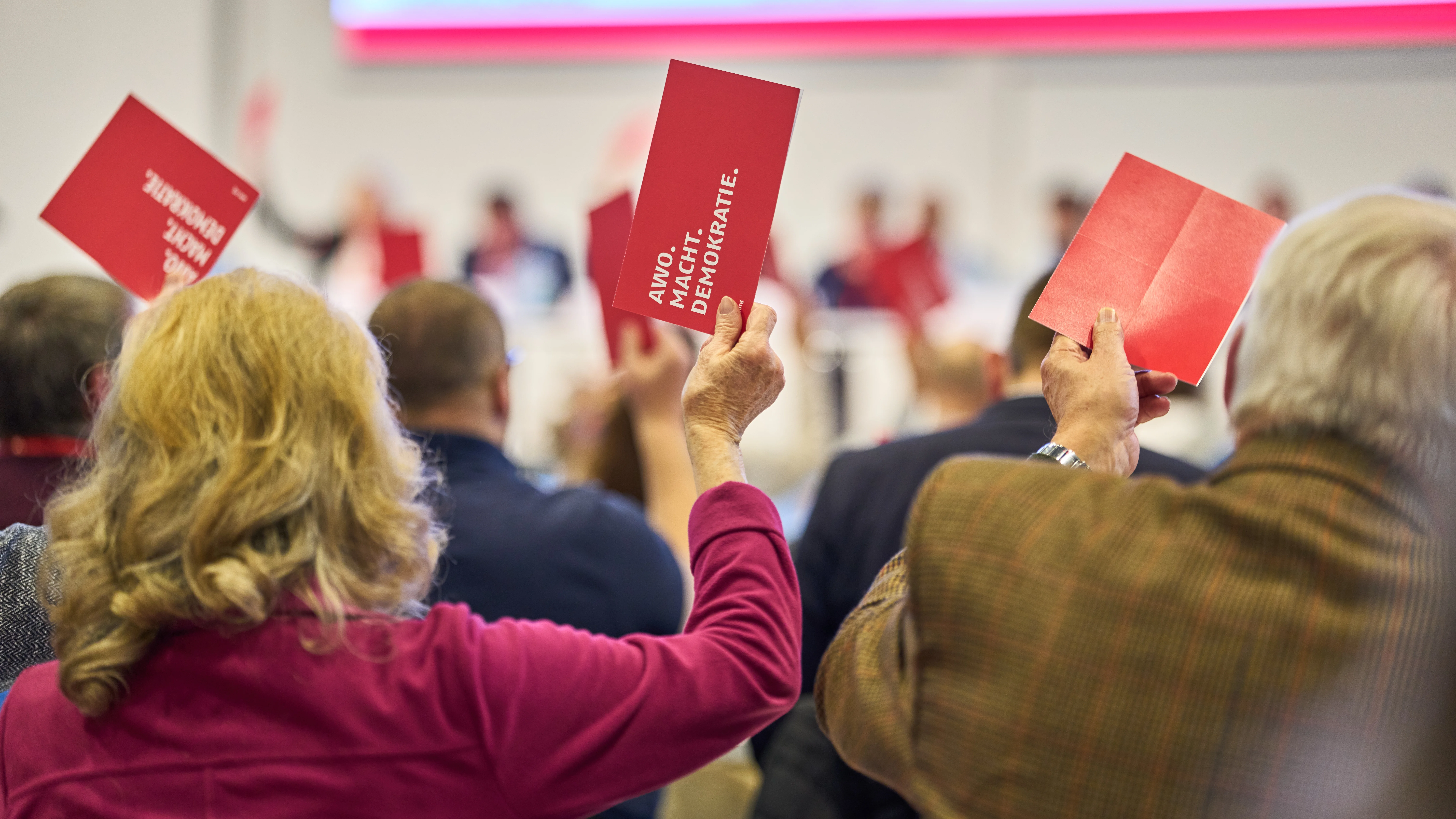 Auf dem Bild ist eine Versammlung oder Konferenz zu sehen, bei der Teilnehmer offensichtlich an einer Abstimmung teilnehmen. Hier sind die Details: Vordergrund: Man sieht die Hinterköpfe und erhobenen Hände von zwei Personen (vermutlich einer Frau mit blonden Locken in einem magenta-farbenen Oberteil und einem Mann mit grauen Haaren und einem braun-grünen Jackett). Abstimmungskarten: Beide halten rote Karten hoch, die als Abstimmungskarten dienen. Text auf den Karten: Auf einer der roten Karten ist Text zu erkennen, der "AWO. MACHT. DEMOKRATIE." lautet. Dies lässt auf eine Veranstaltung der AWO (Arbeiterwohlfahrt) schließen. Hintergrund: Weitere Teilnehmer im Saal heben ebenfalls rote Karten. Im unscharfen Hintergrund ist eine helle Projektionsfläche oder ein Banner zu sehen. Zusammenfassend handelt es sich um eine Szene einer demokratischen Abstimmung bei einer Veranstaltung der AWO.