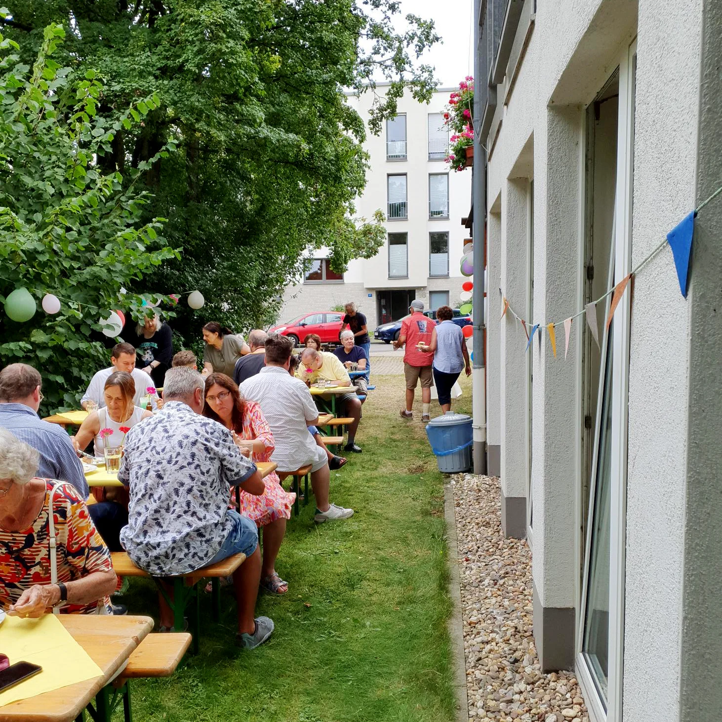 Das Foto zeigt eine fröhliche Zusammenkunft in einem Garten mitten in einem Wohngebiet. Mehrere Tische sind aufgestellt, an denen Menschen sitzen, essen und sich unterhalten. Die Umgebung umfasst ein Gebäude auf der rechten Seite mit Fenstern und Pflanzen auf den Fensterbänken sowie ein weiteres Gebäude im Hintergrund. Dekorationen wie Luftballons und Girlanden deuten darauf hin, dass es sich um eine festliche oder gemeinschaftliche Veranstaltung handelt.