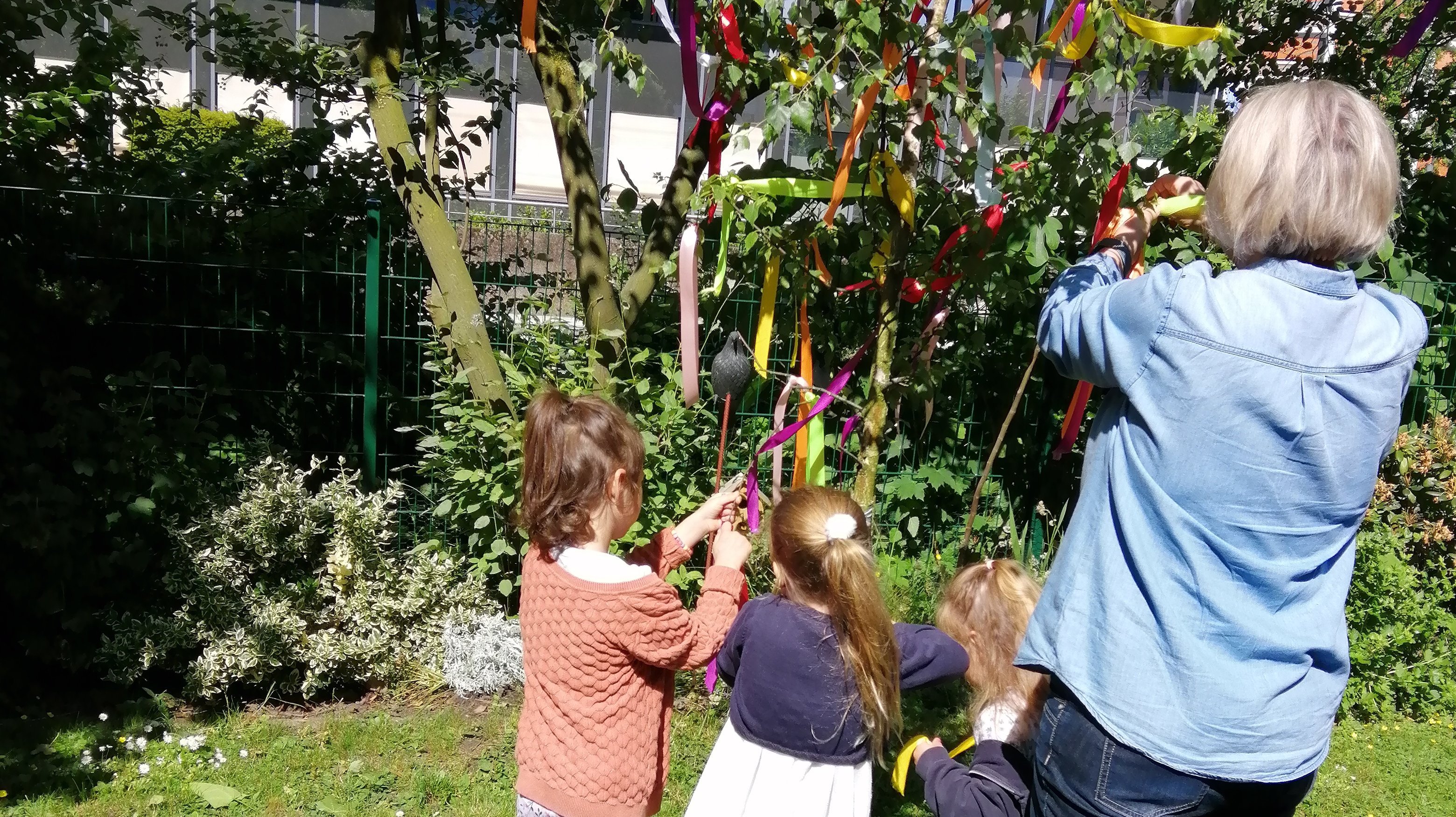 Dieses Bild zeigt eine Erzieherin wie Sie gemeinsam mit den Kindern den Maibaum abschmückt.