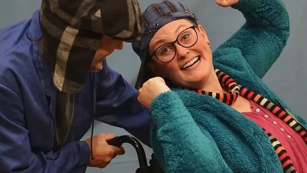 Auf dem Foto sind zwei Erwachsene in einer freudigen oder spielerischen Interaktion zu sehen, möglicherweise im Rahmen einer Aufführung oder eines Sketche. Die Person rechts trägt eine türkis-blaue, flauschige Jacke, ein pinkes Shirt und einen gestreiften Schal. Sie hat eine Brille auf und trägt eine Baseballkappe verkehrt herum auf dem Kopf. Ihr Ausdruck ist ausgelassen und lachend, die Faust ist erhoben. Die Person links trägt eine blaue Arbeitskleidung oder einen Overall und eine karierte Mütze mit Ohrenklappen. Man sieht nur einen Teil ihres Gesichts, das sich der lachenden Person zuneigt. Es scheint, als würde die linke Person einen Griff (möglicherweise von einem Rollstuhl oder einem Requisit) halten, den die rechte Person leicht nach hinten gelehnt hält.