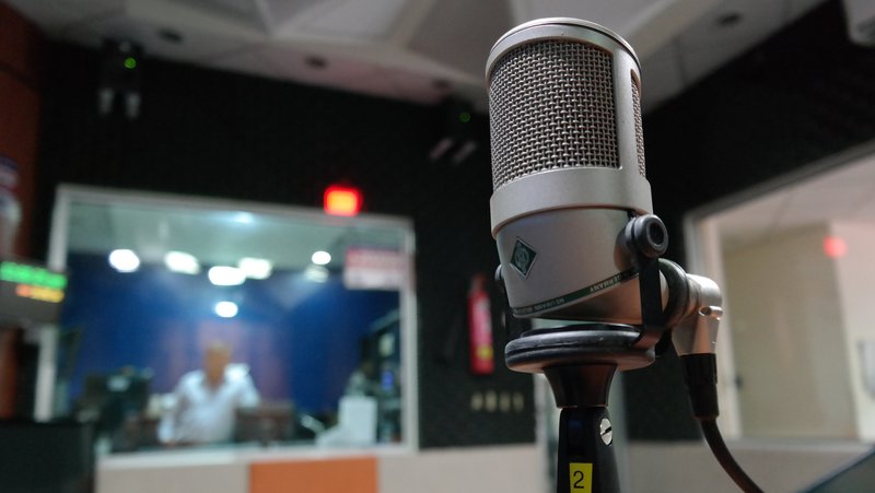 Im Vordergrund ist ein großes Studio‑Mikrofon auf einem Stativ zu sehen; im Hintergrund erkennt man unscharf einen Radiostudio‑Raum mit Person hinter einer Glasscheibe und verschiedenen Geräten.